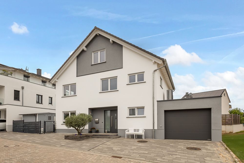 Modernes, weißes Wohnhaus mit grauer Garage, Baum und Bank vor dem Eingang.