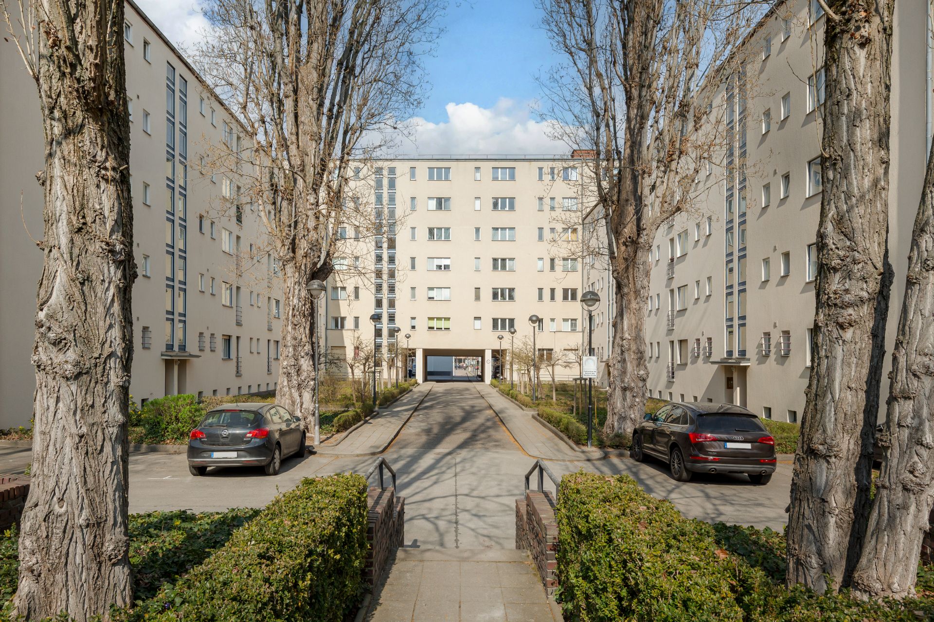 Eingangsbereich eines Wohnblocks mit Bäumen, Autos und Gehweg.