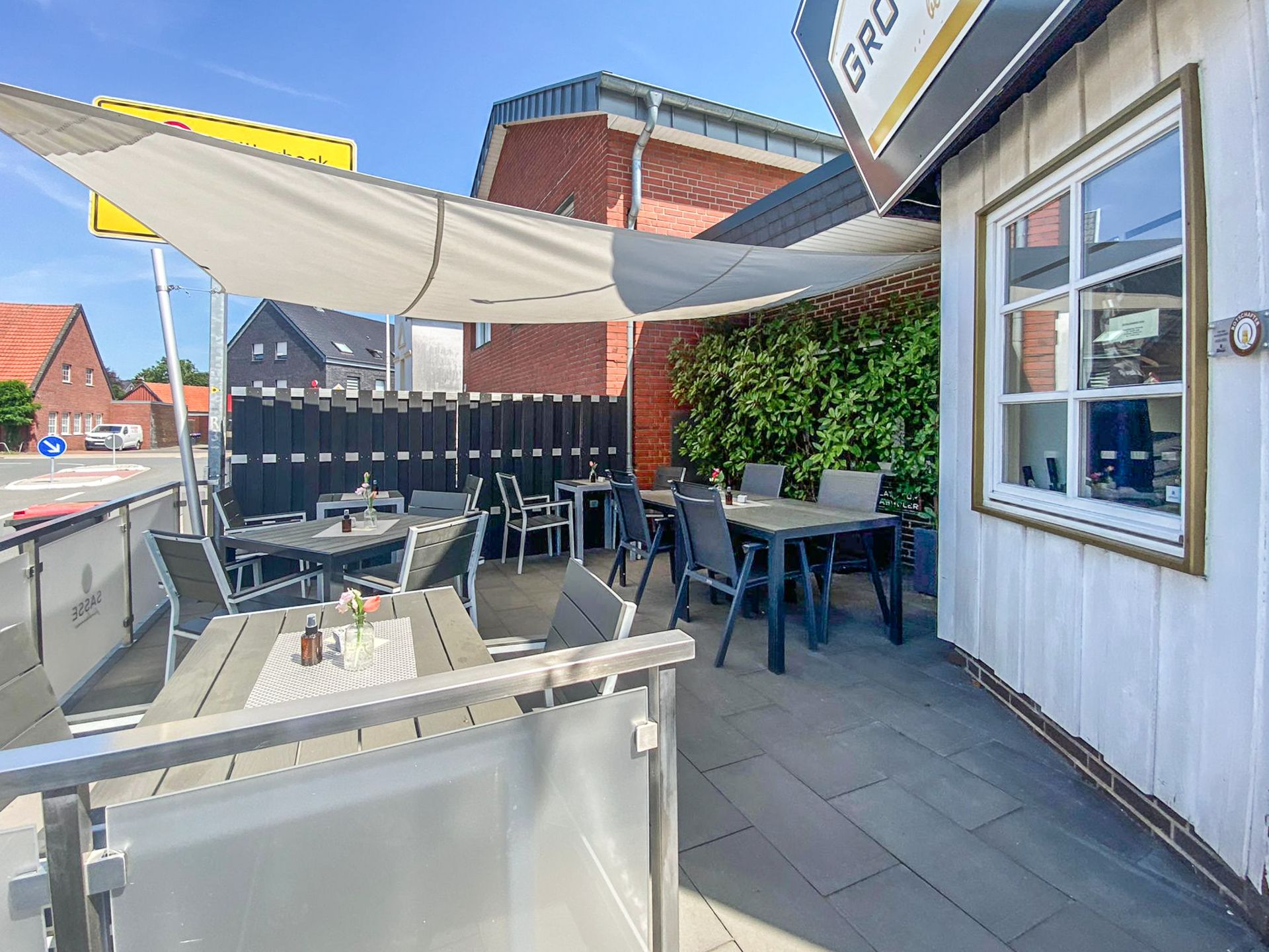 Terrasse mit mehreren Tischen und Stühlen, Sonnensegel und Pflanzenwand, bei Tageslicht.