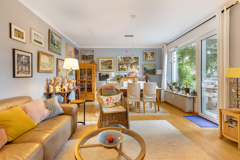 Helles Wohnzimmer mit Sofa, Esstisch, vielen Bildern an den Wänden und großen Fenstern mit Blick ins Grüne.
