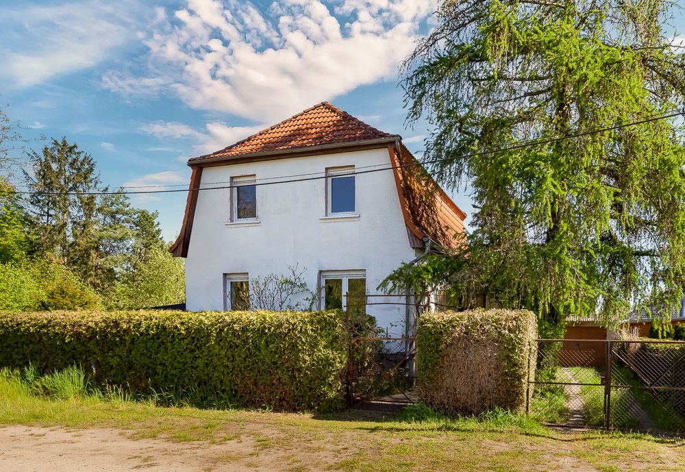 Weißes Einfamilienhaus mit rotem Dach, umgeben von Hecken und Bäumen, kleiner Garten vor dem Haus.