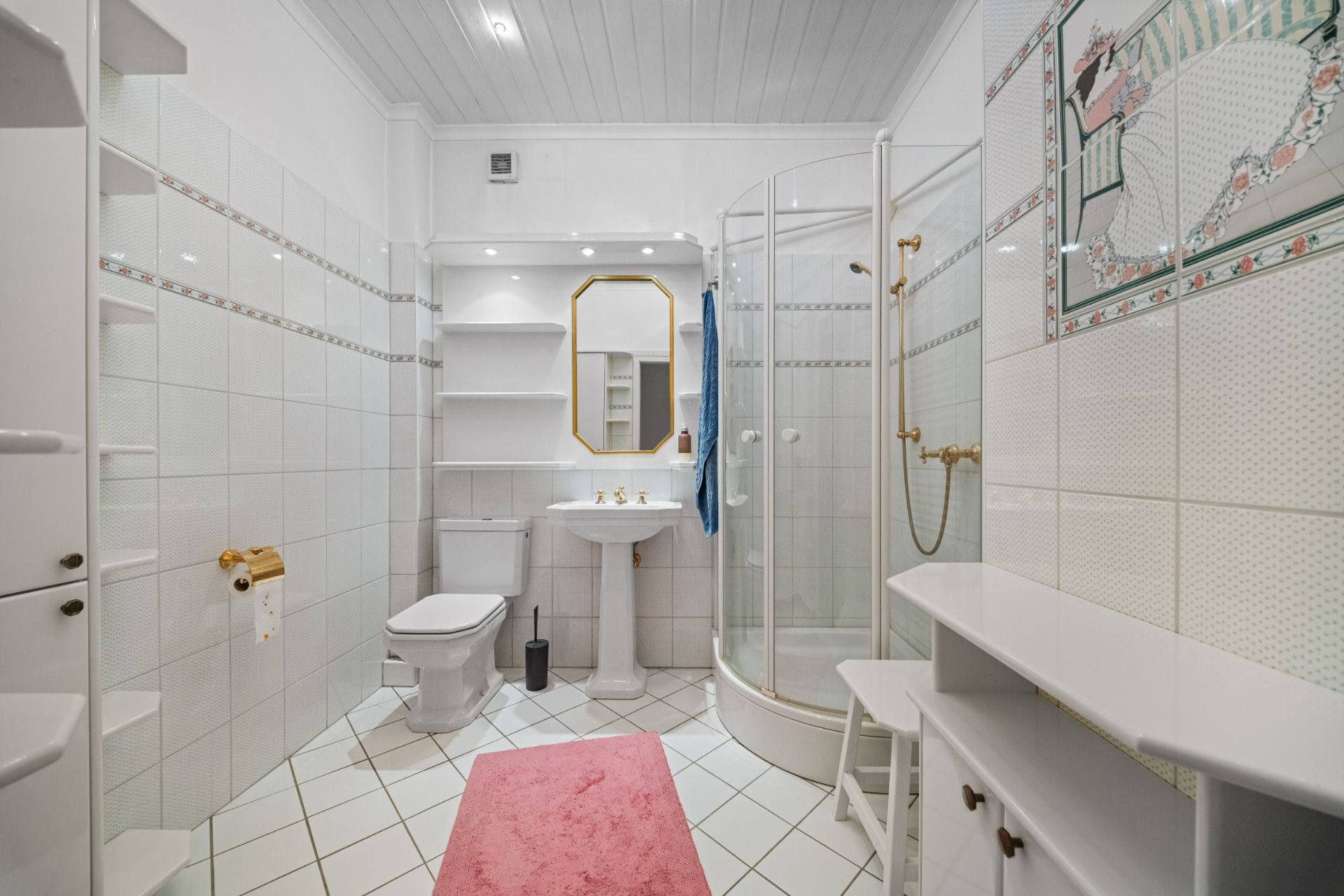 Helles Badezimmer mit Dusche, WC, Waschbecken, Regal und rosa Teppich auf weißen Fliesen.