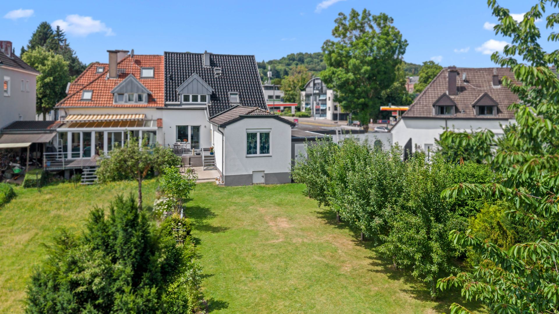 Haus mit weißer Fassade, großem Garten und Bäumen, im Hintergrund weitere Wohnhäuser.