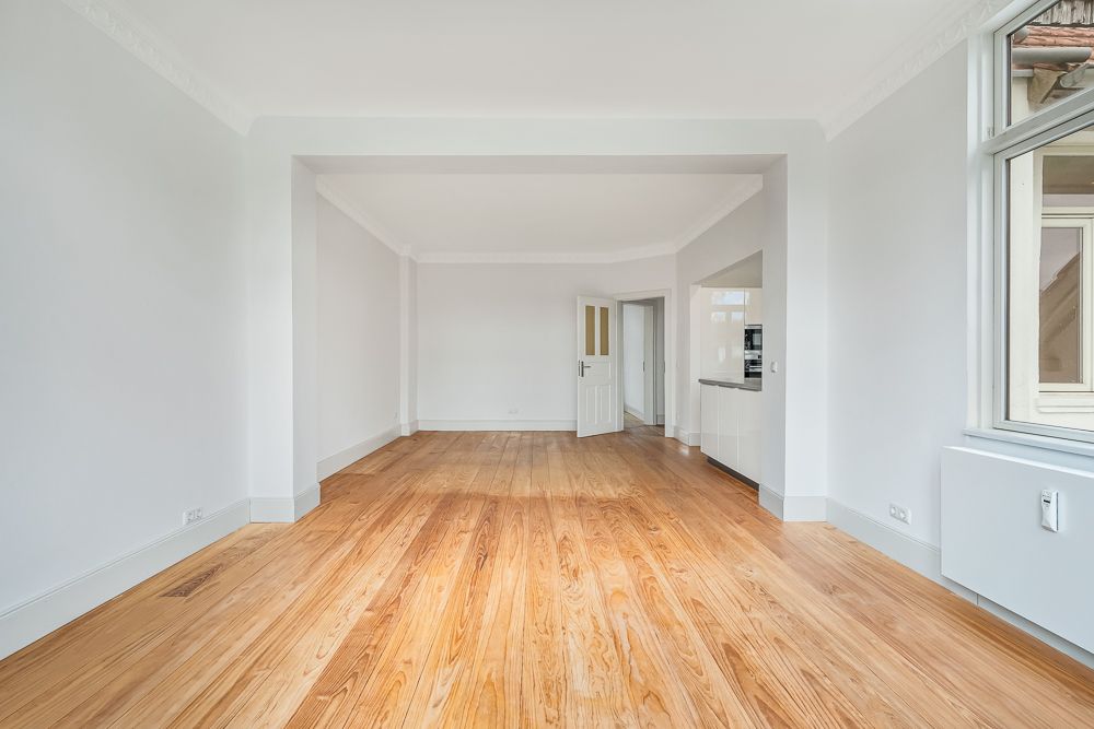Helles Wohnzimmer mit Holzfußboden, weißen Wänden, Fenstern und einer offenen Tür zur Küche.