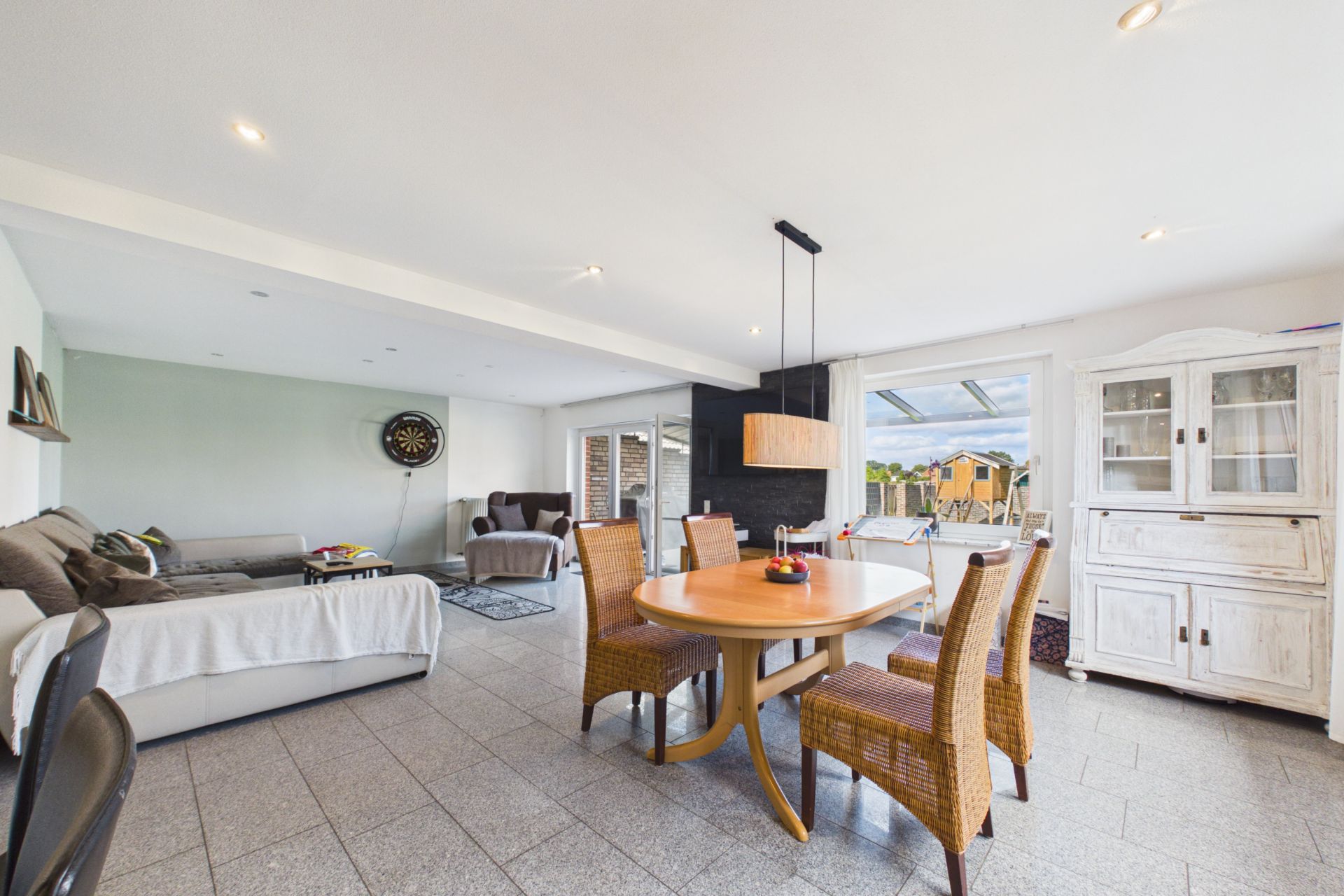 Helles Wohnzimmer mit Esstisch, vier Stühlen, Sofa, Vitrine und großen Fenstern.