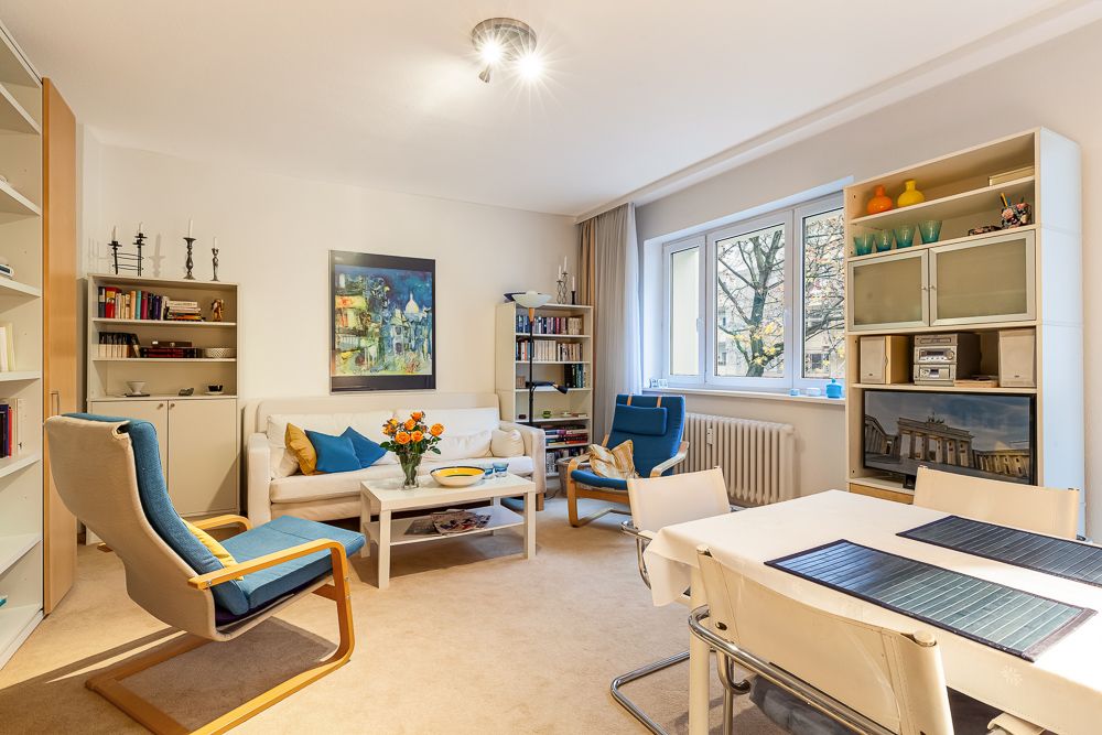 Helles Wohnzimmer mit Sofa, Esstisch, Regalen, Sessel und einem Fenster mit Blick auf Bäume.