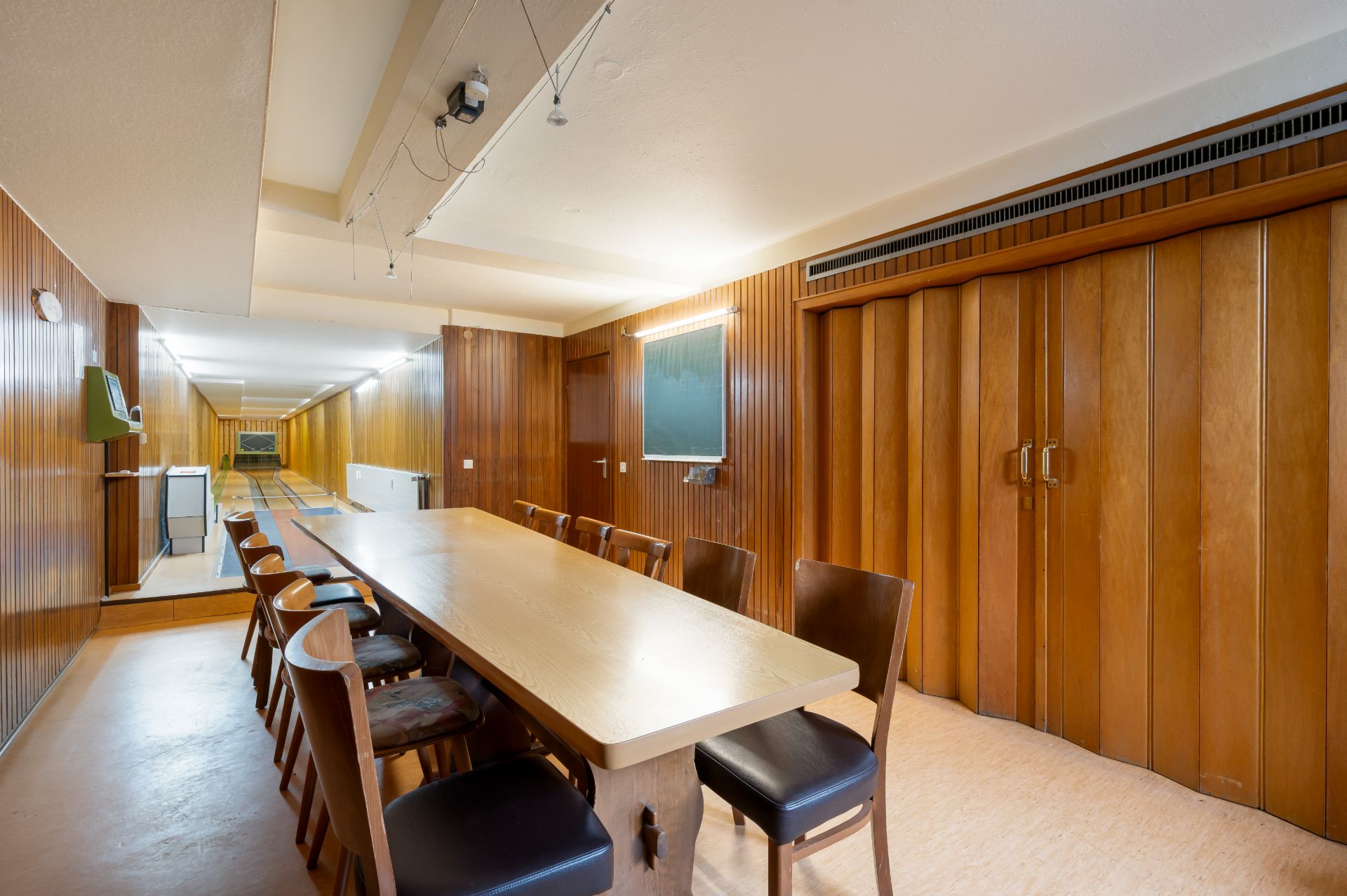 Langer Holztisch mit mehreren Stühlen in einem Raum mit Holzverkleidung und Tafel an der Wand.