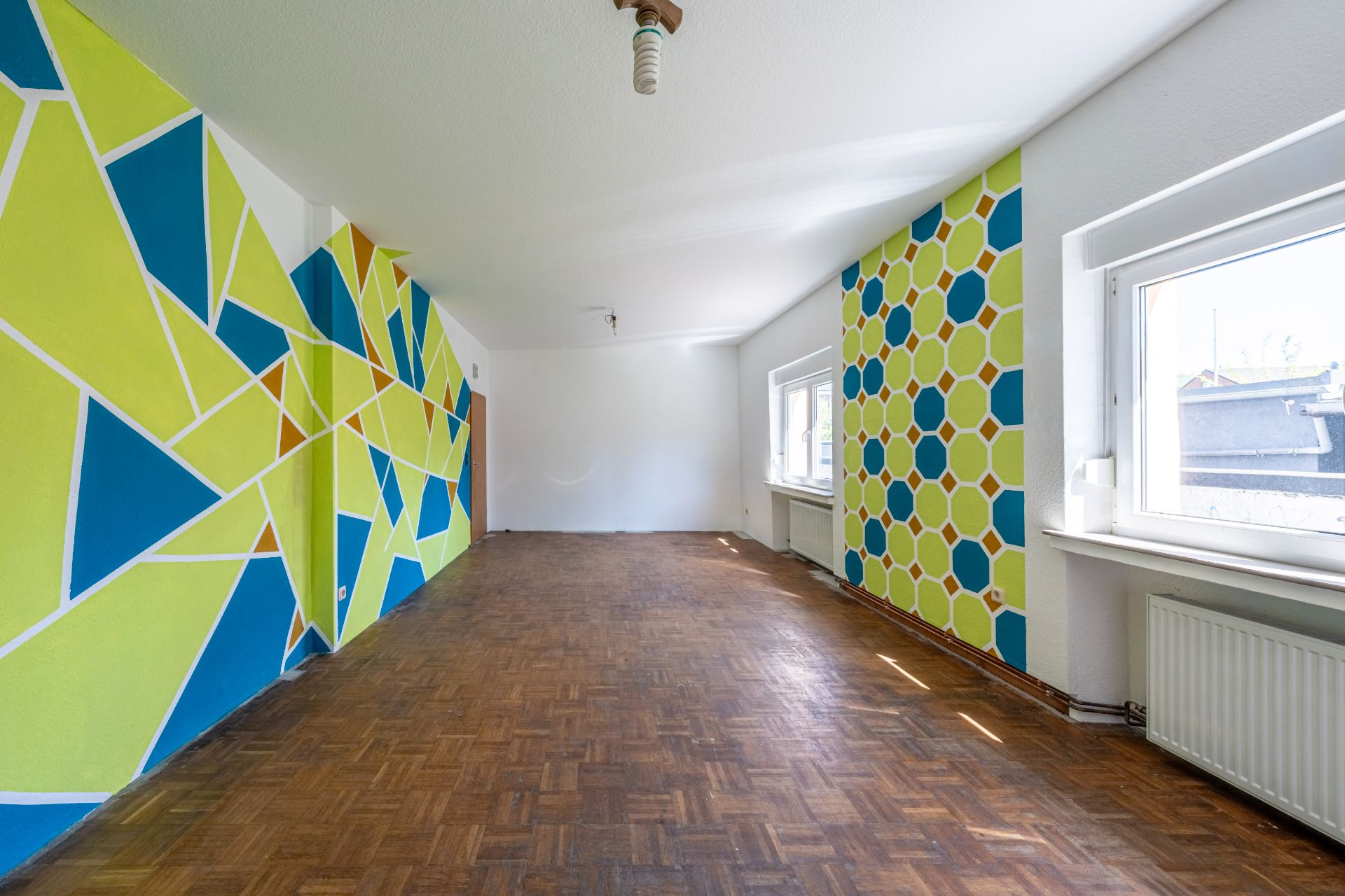 Leeres Zimmer mit bunten, geometrischen Mustern an den Wänden und Parkettboden.