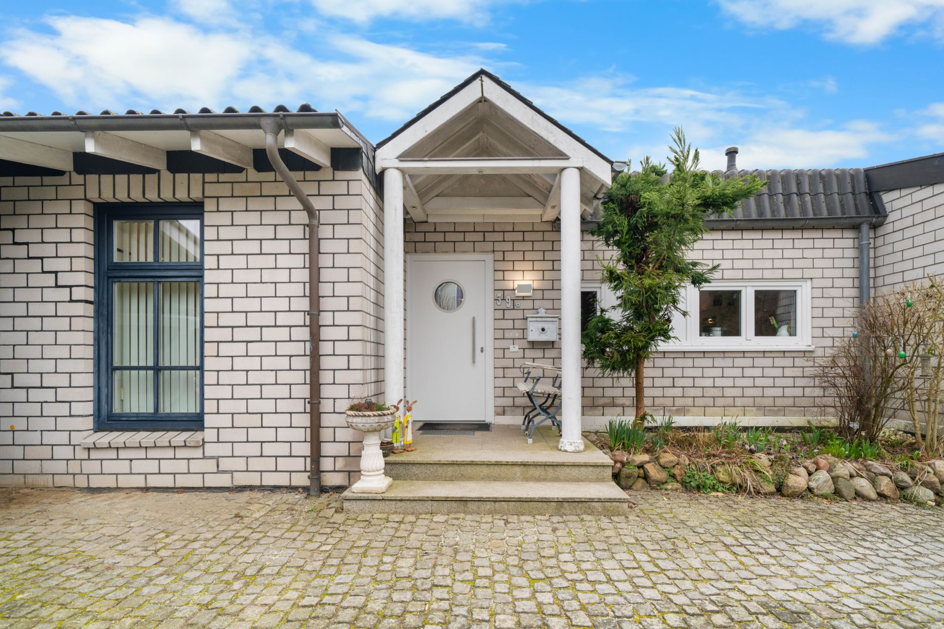 Eingang eines Einfamilienhauses mit weißer Fassade, überdachter Tür und kleinem Gartenbereich.