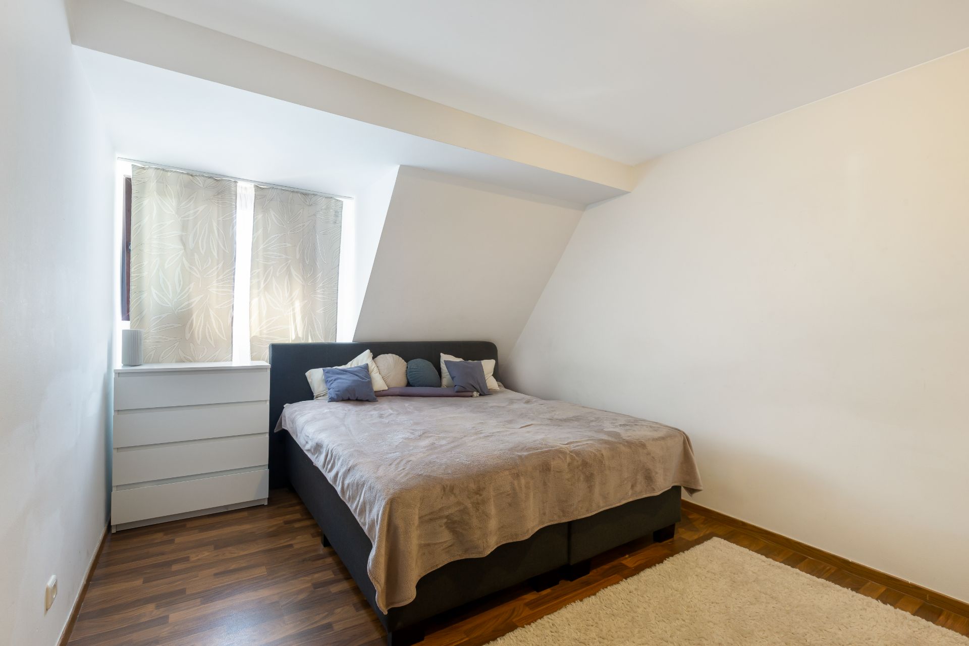 Helles Schlafzimmer mit Doppelbett, mehreren Kissen, Kommode und Teppich auf Holzfußboden.