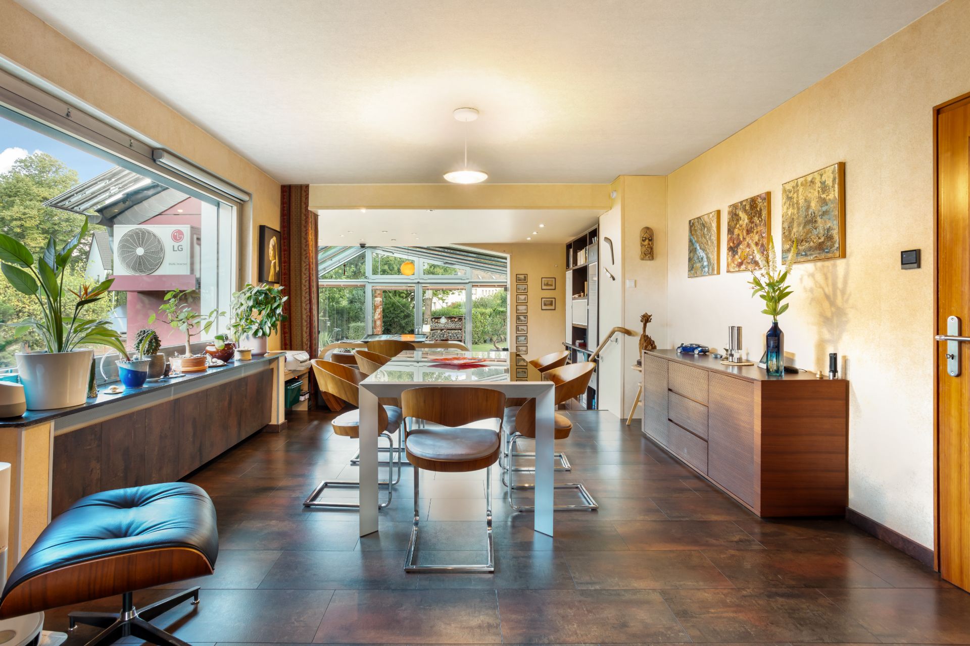 Helles Wohnzimmer mit großem Fenster, Esstisch, Pflanzen und Holzkommode.