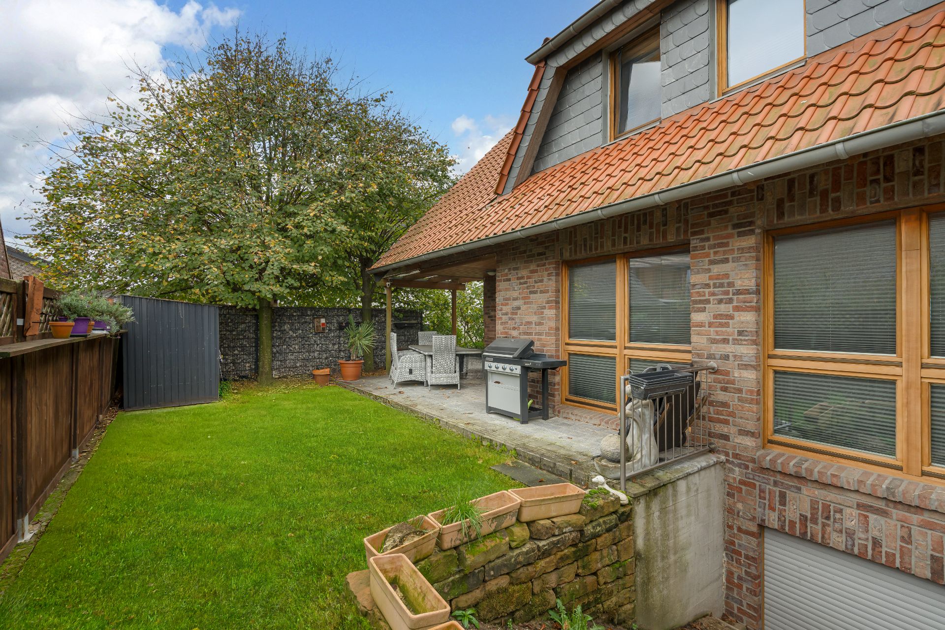 Garten mit Terrasse, Grill, Sitzmöbeln, Baum und Hochbeeten an einem Wohnhaus.