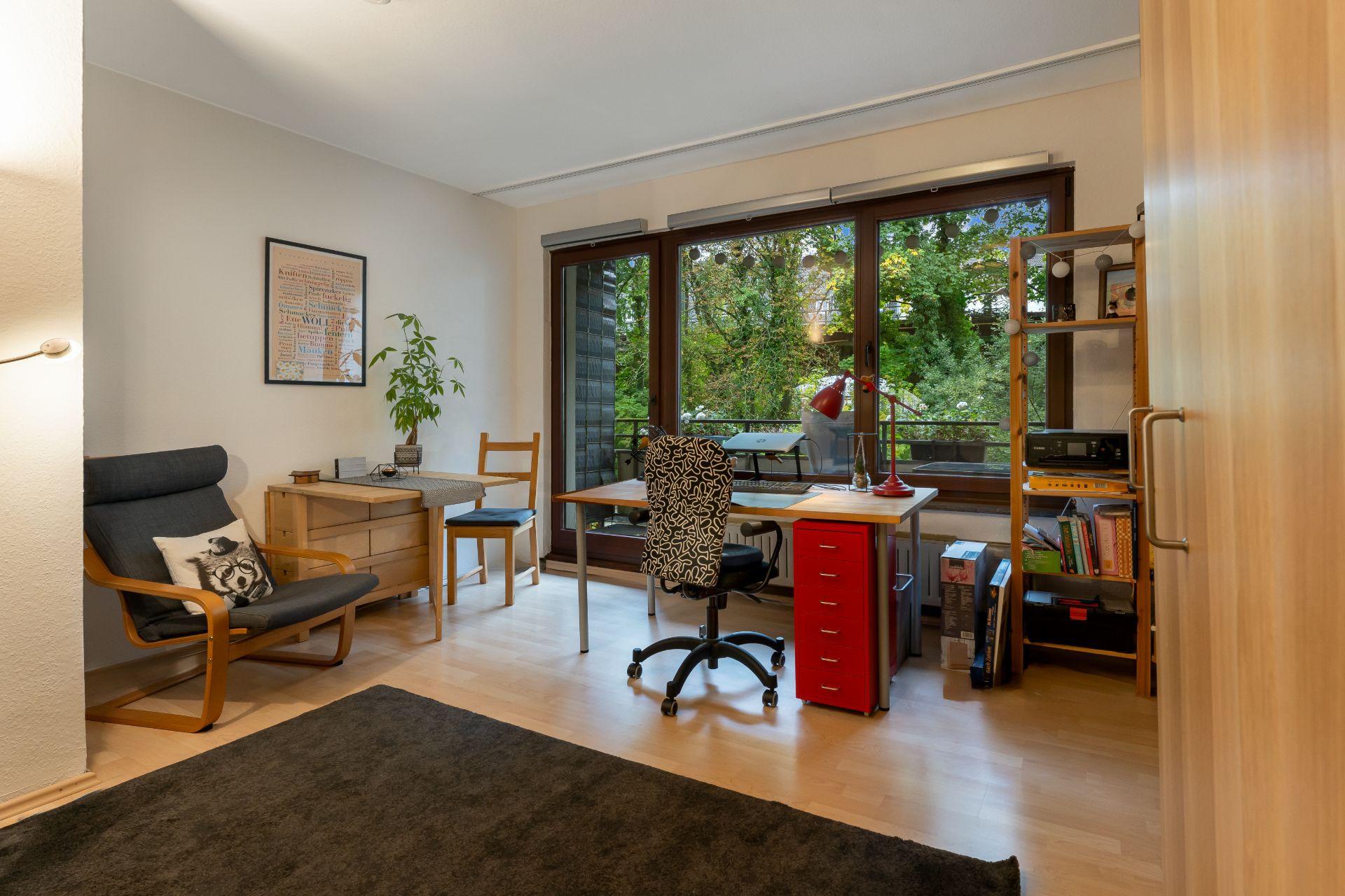 Helles Arbeitszimmer mit Schreibtisch, Stuhl, Regalen und großem Fenster zum Garten.