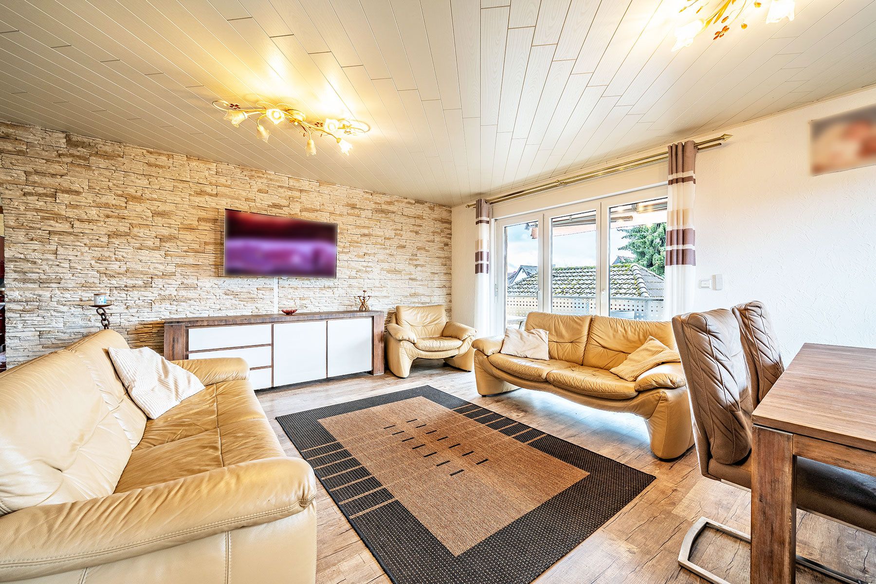 Helles Wohnzimmer mit beigen Ledersofas, Teppich und großem Fenster, Steinwand und Fernsehgerät.