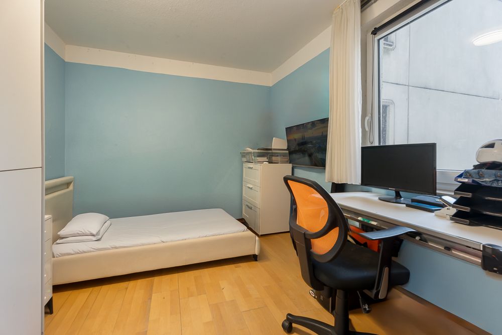 Kleines Zimmer mit Einzelbett, Schreibtisch, Stuhl und Computer neben einem Fenster.