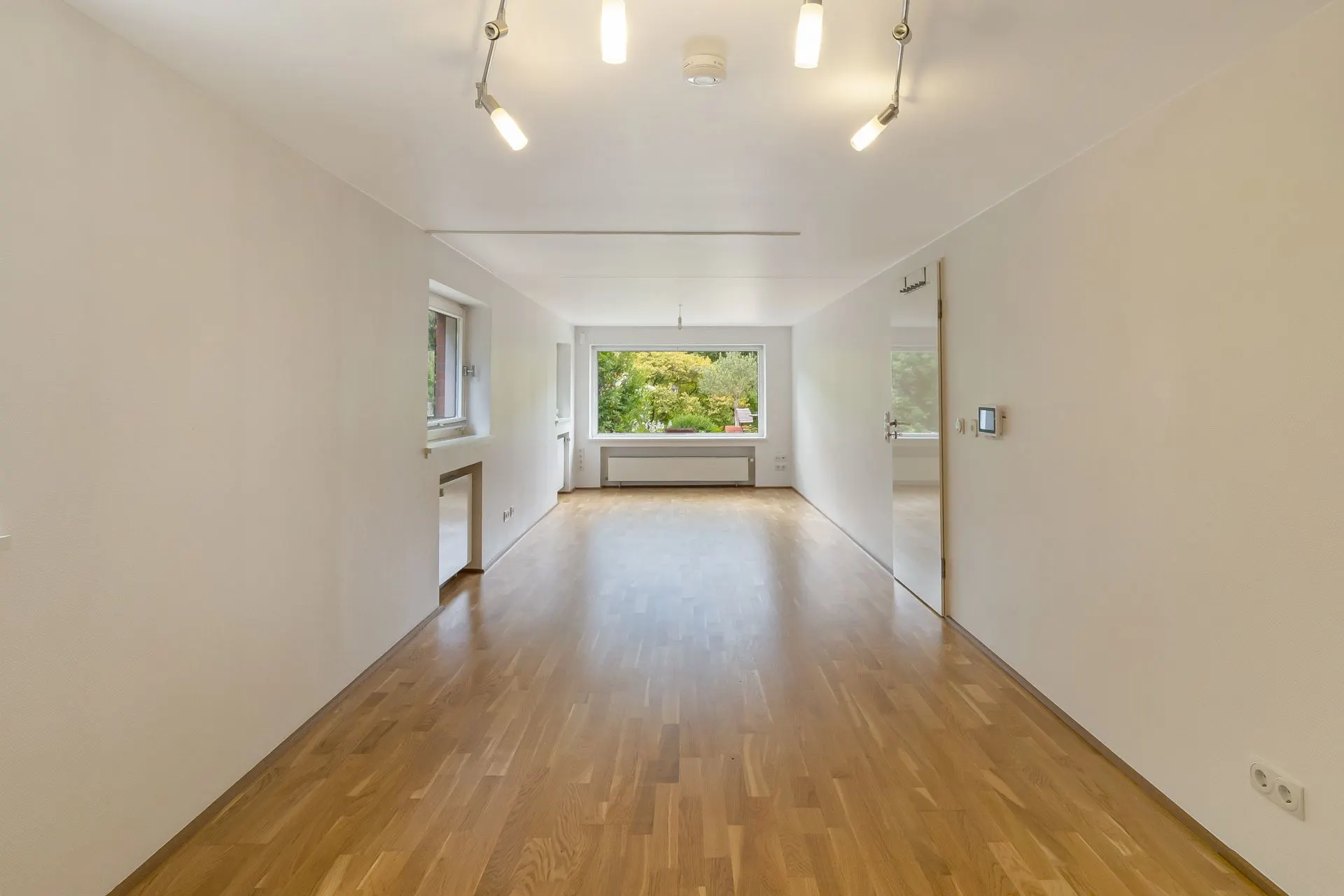 Helles, leeres Wohnzimmer mit Holzfußboden, weißen Wänden und großem Fenster mit Blick ins Grüne.