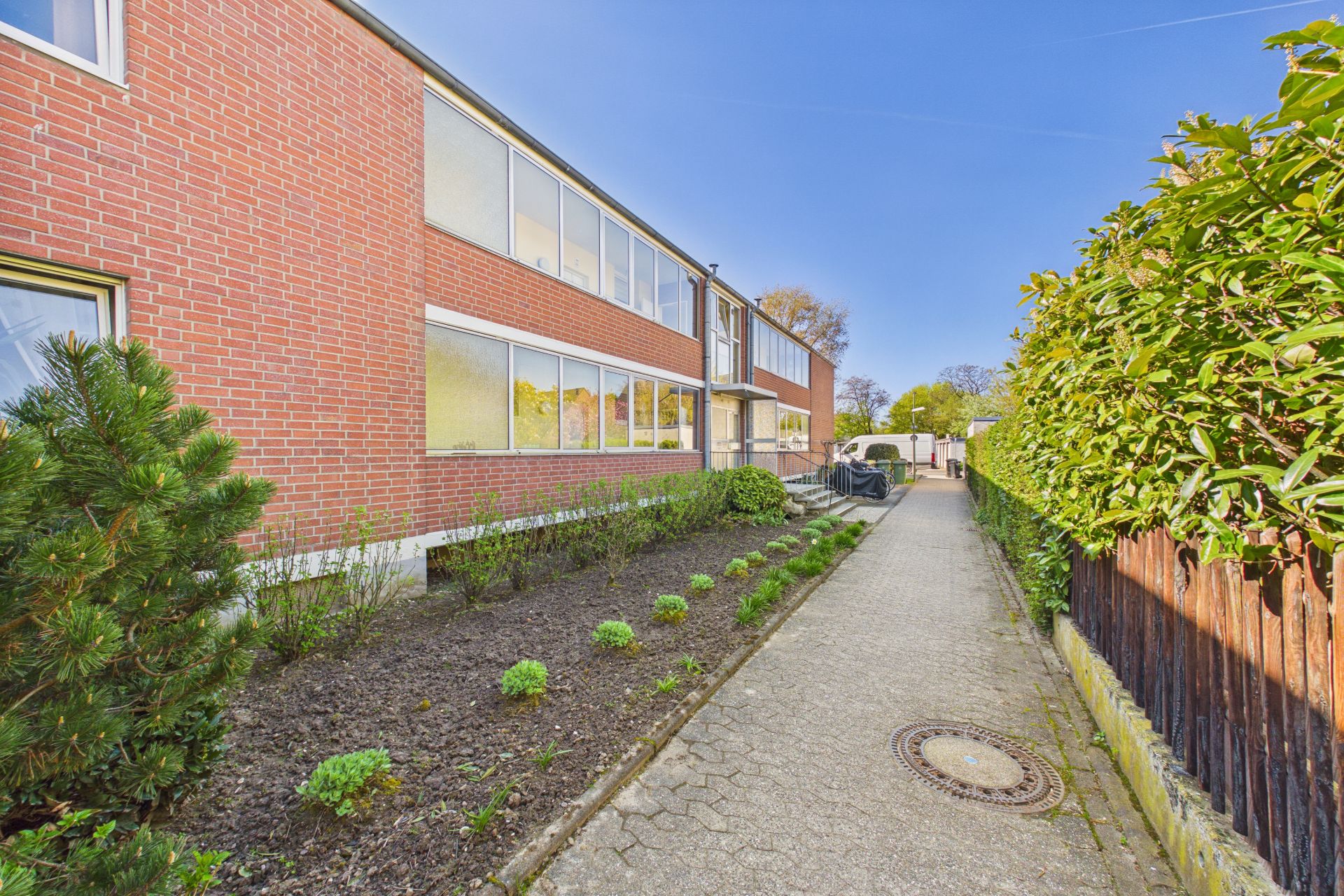 Gepflasterter Weg neben einem zweistöckigen roten Backsteingebäude mit Garten und Hecke.