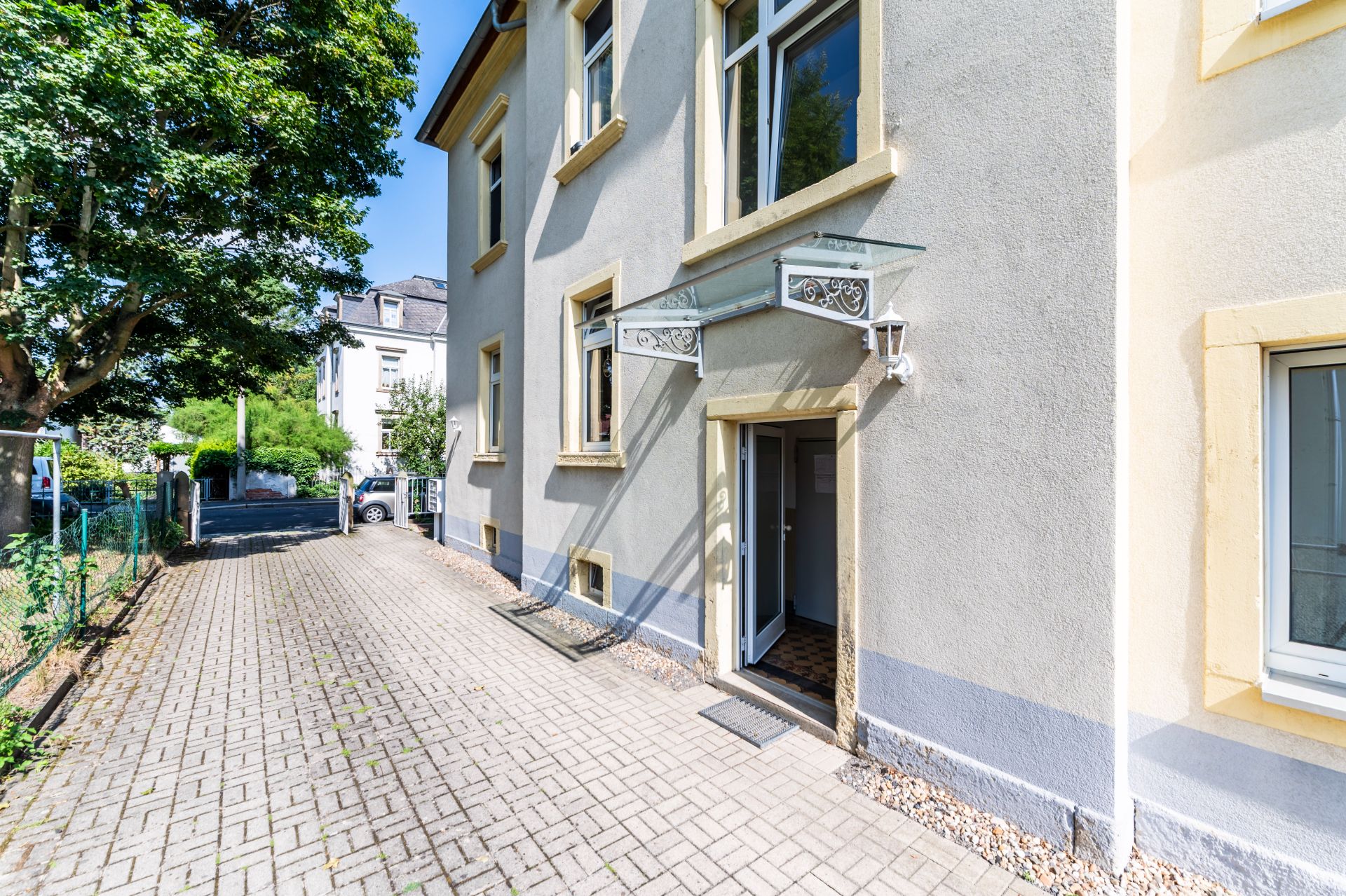 Eingang eines Wohnhauses mit offen stehender Tür und kleinem Glasvordach, daneben gepflasterter Weg.