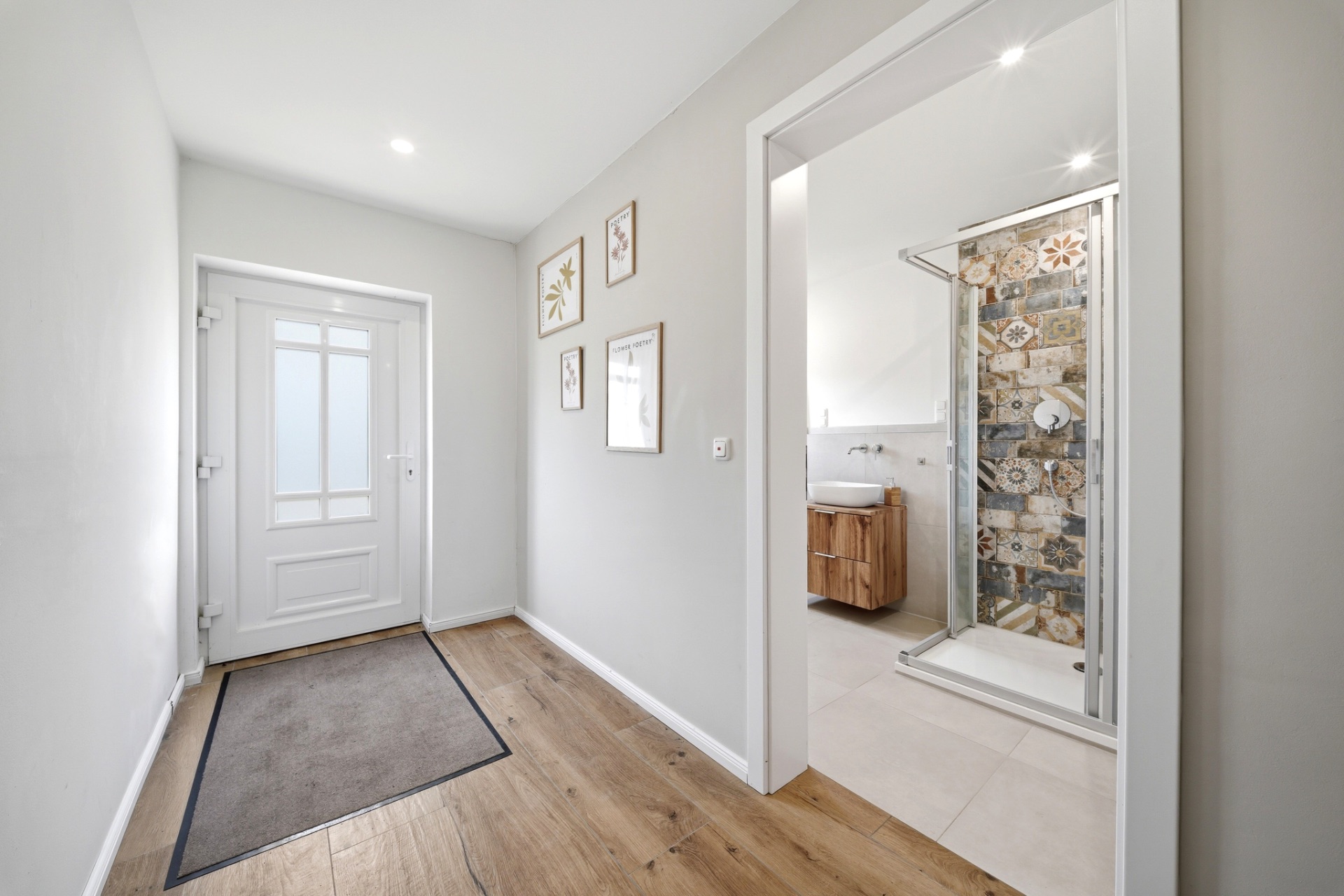 Heller Flur mit Holzboden, Bilder an der Wand und Blick ins Badezimmer mit Dusche.