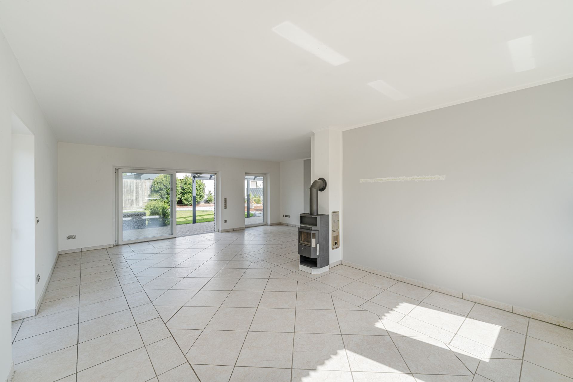 Heller, leerer Wohnraum mit Fliesenboden, Kaminofen und großen Fenstern zum Garten.