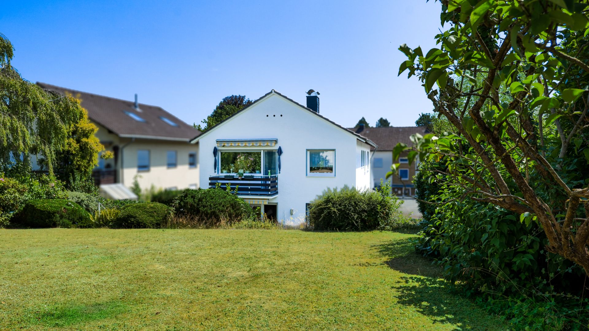Weißes Einfamilienhaus mit Balkon, Garten und Bäumen im Vordergrund an einem sonnigen Tag.