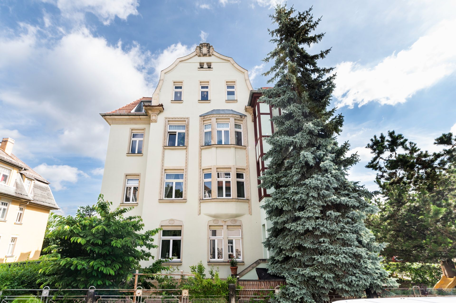 Mehrstöckiges Wohnhaus mit vielen Fenstern, großen Bäumen und Garten vor dem Gebäude.