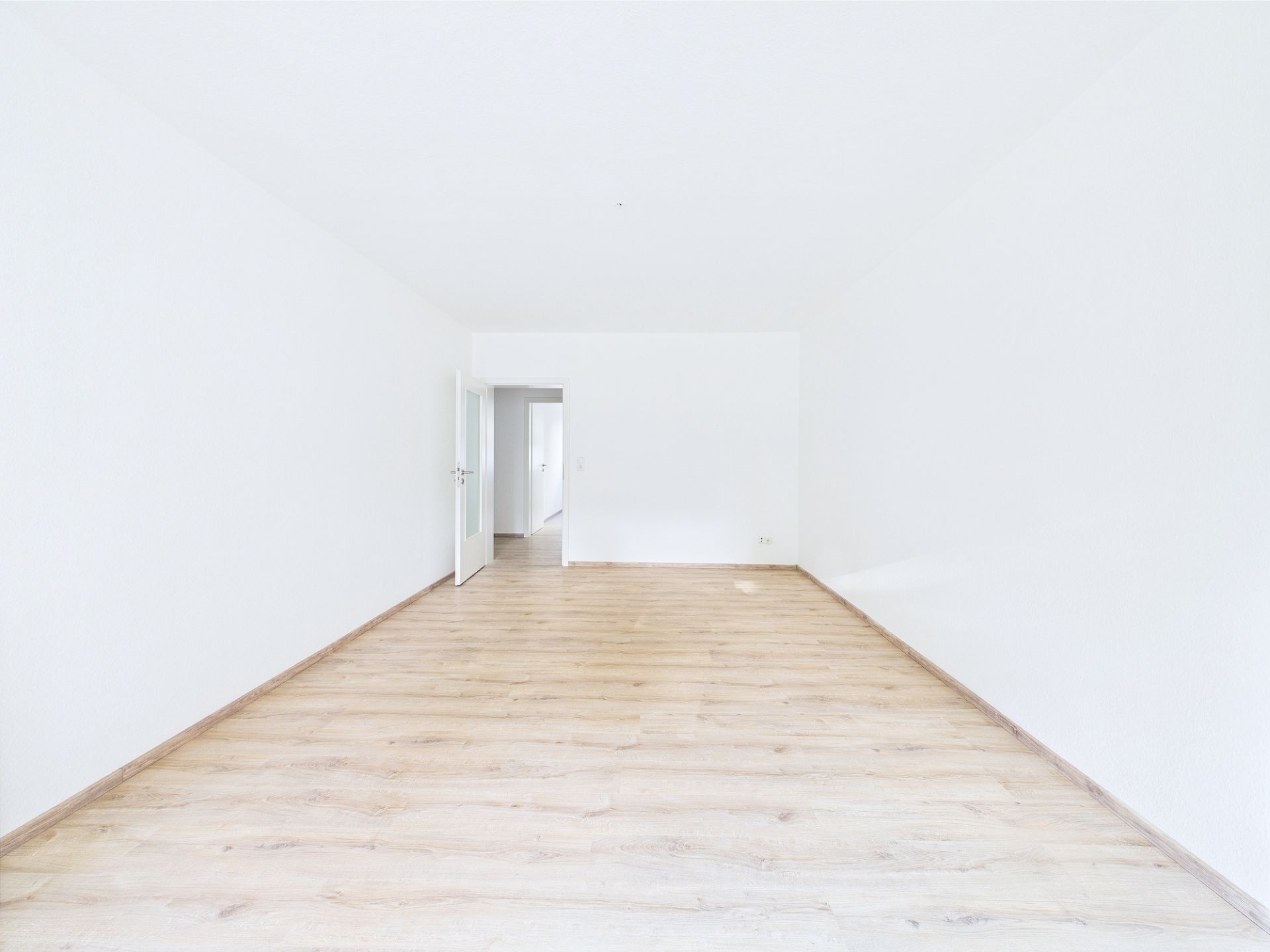 Leerer, heller Raum mit Holzfußboden und weißen Wänden, Tür führt in einen Nebenraum.