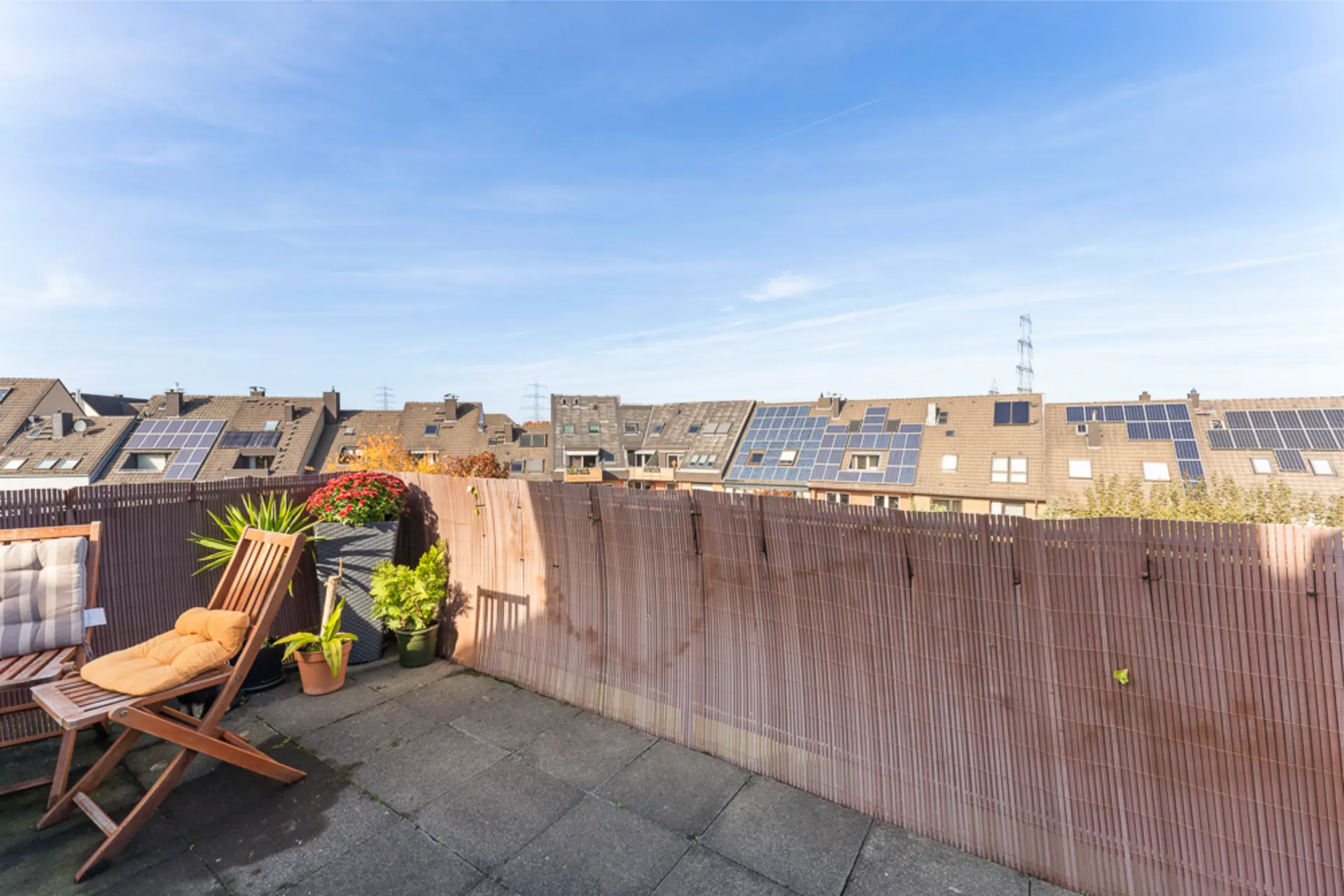 Sonnige Dachterrasse mit Holzstuhl, Pflanzen und Blick auf Häuser mit Solarpaneelen.