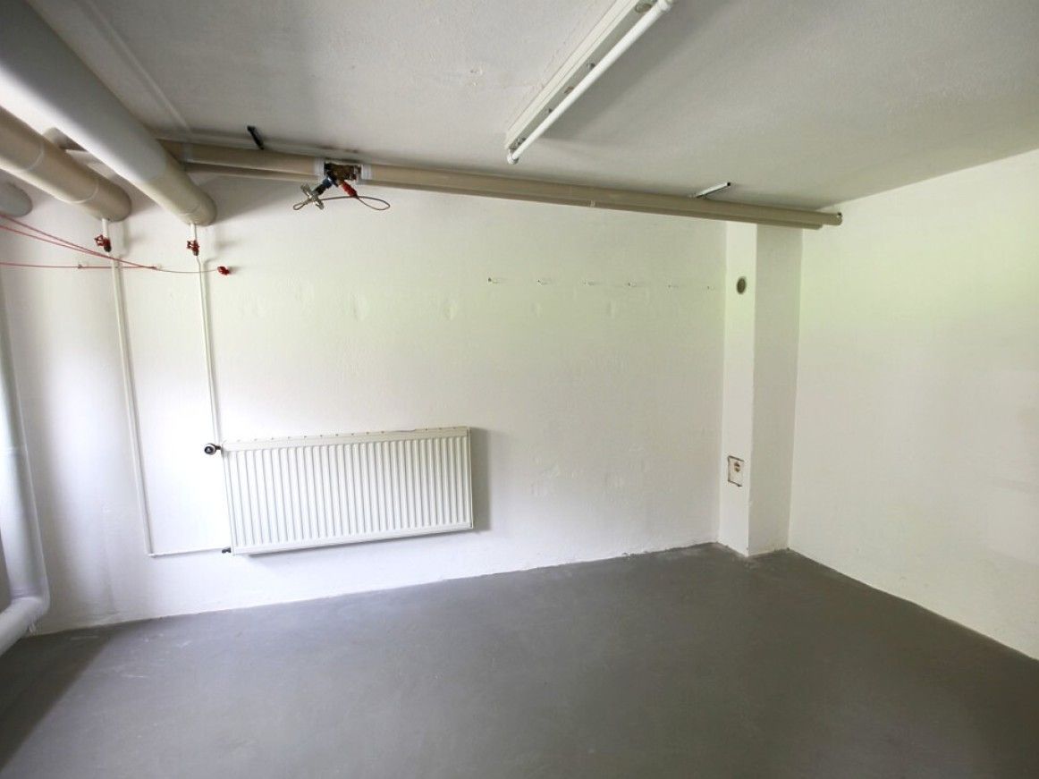 Leerer Kellerraum mit weiß gestrichenen Wänden, Heizkörper und sichtbaren Rohrleitungen.