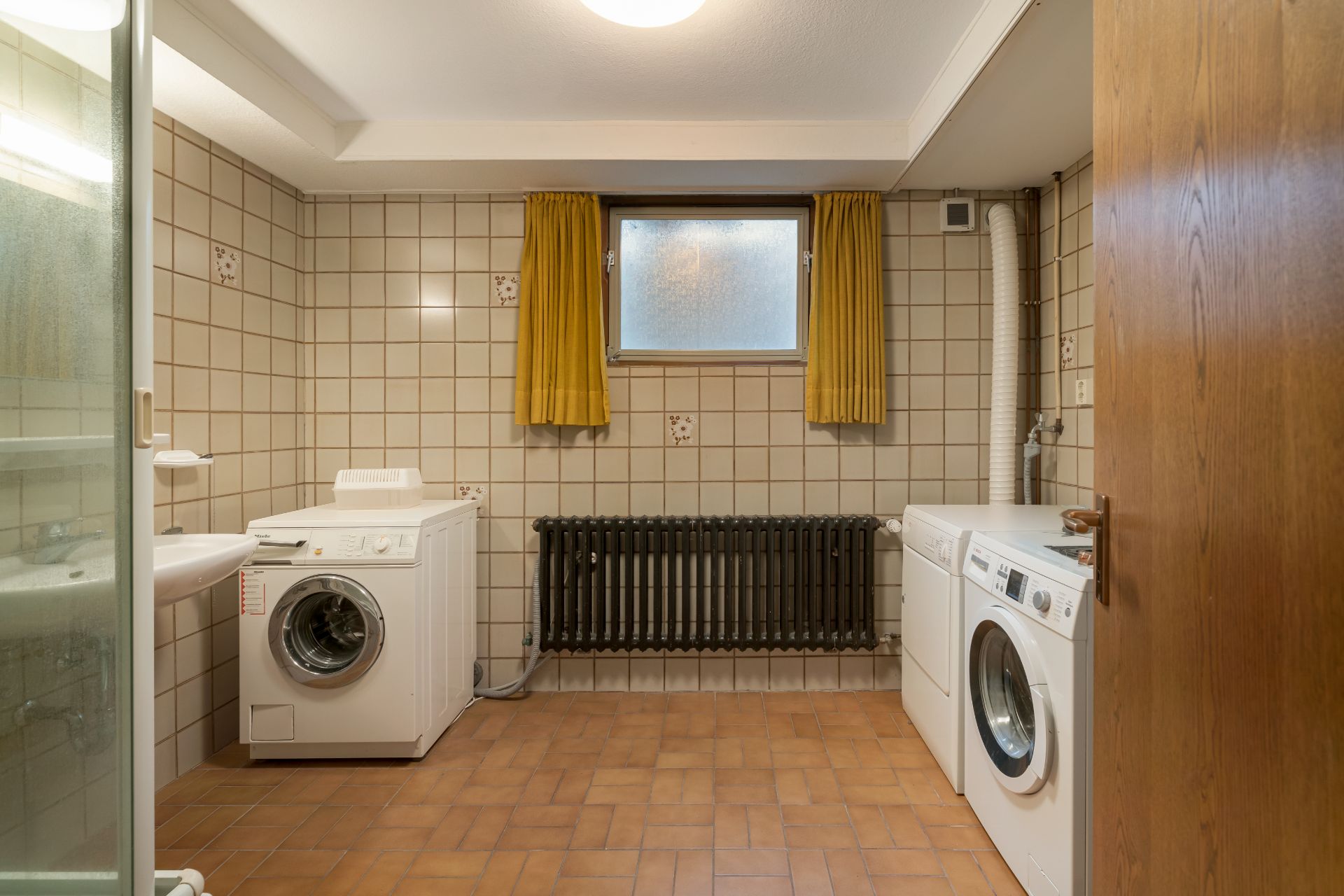 Waschküche mit zwei Waschmaschinen, Heizkörper, kleinem Fenster und gelben Vorhängen.