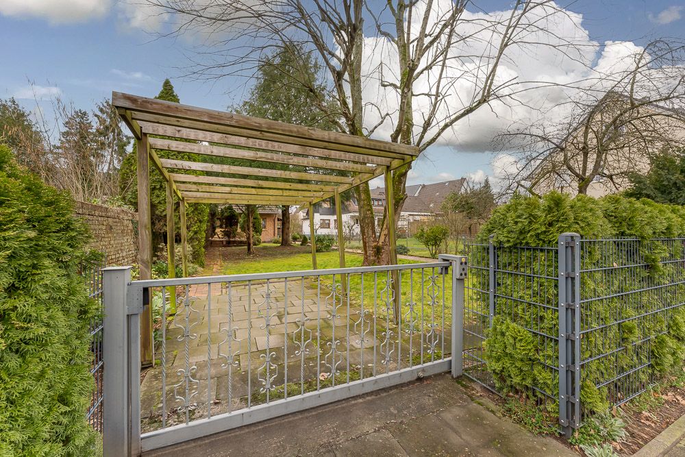Gartentor mit Metallzaun und Holzpergola, dahinter Rasen und Bäume.