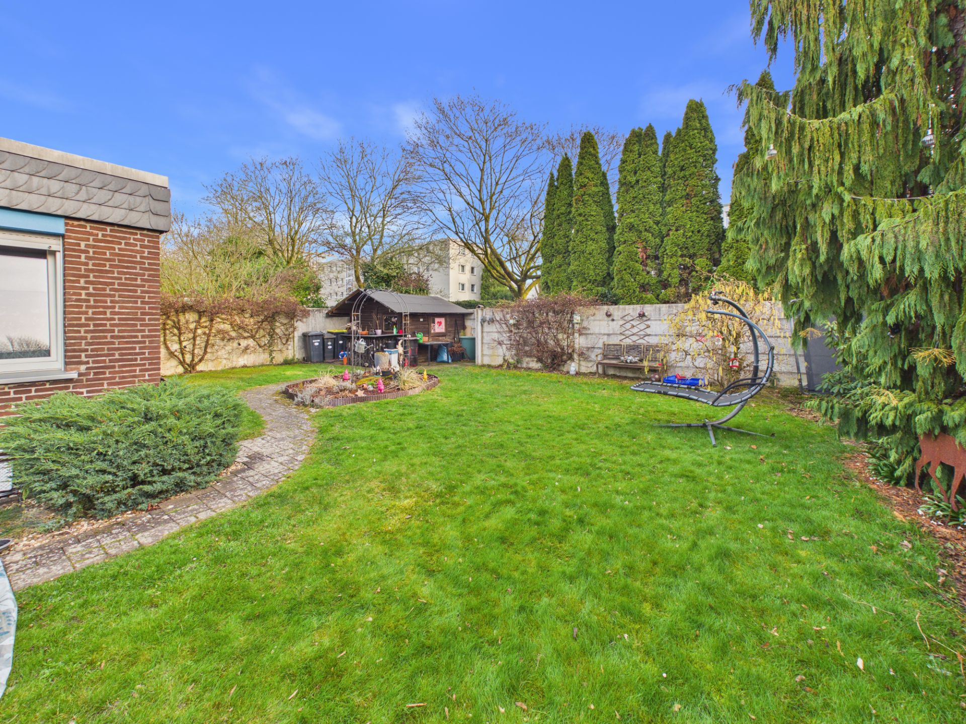 Grüner Garten mit Rasen, Gartenhaus, Schaukelstuhl und Bäumen an einer Steinmauer.
