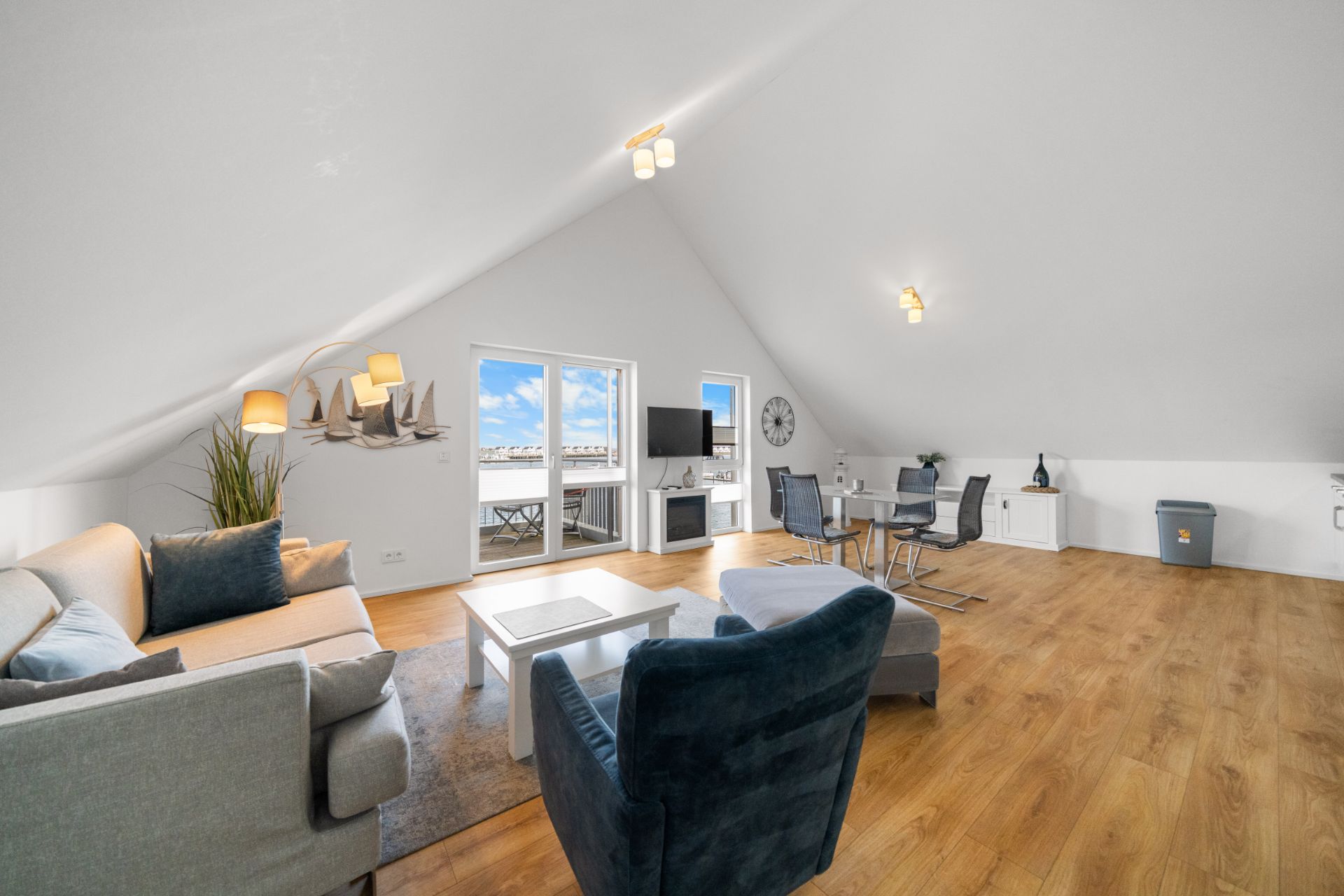 Modernes Wohnzimmer mit Dachschräge, Sofa, Sessel, Esstisch und TV, Holzboden und Zugang zum Balkon.