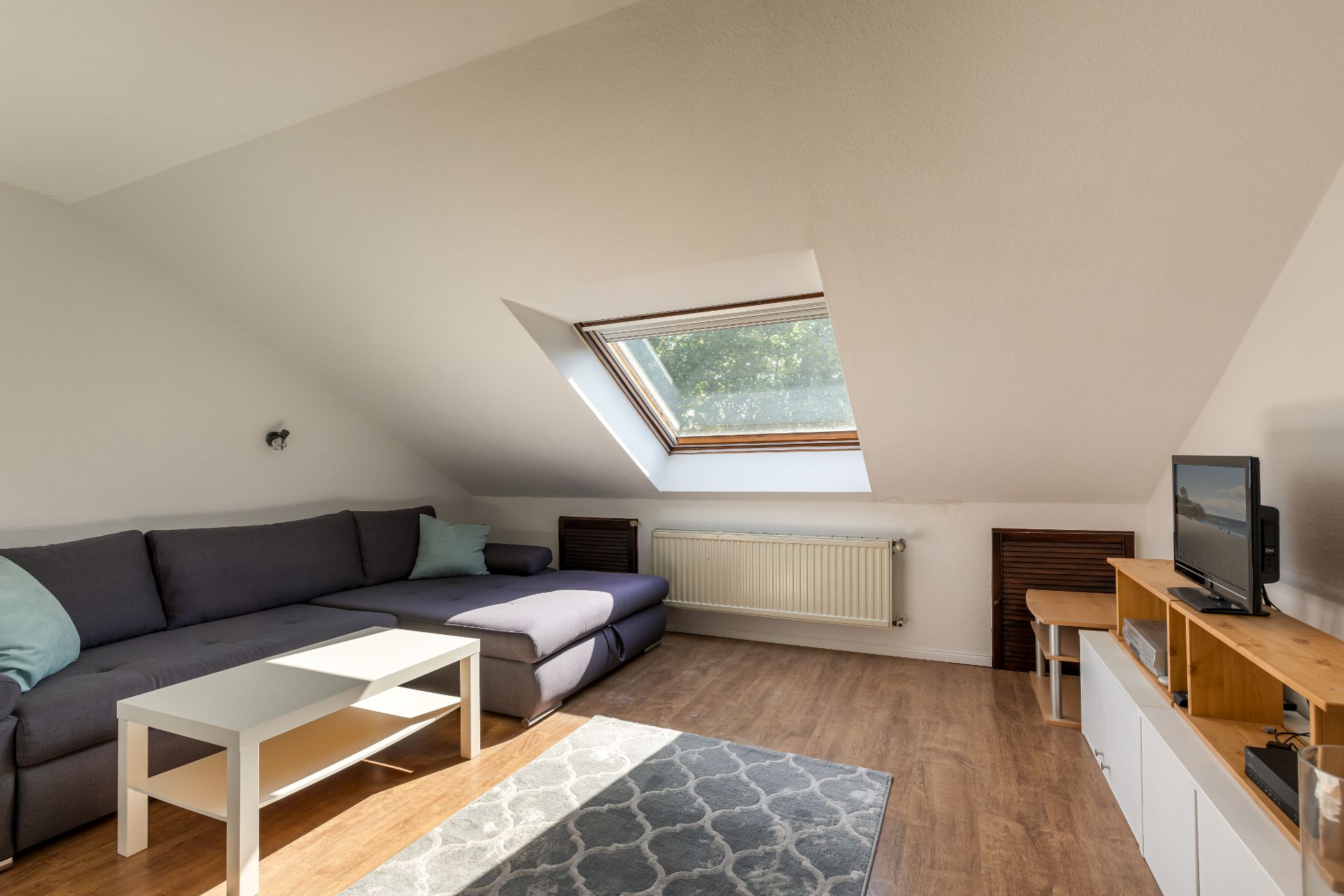Wohnzimmer mit grauem Sofa, Couchtisch, Teppich und TV unter Dachschräge mit großem Fenster.