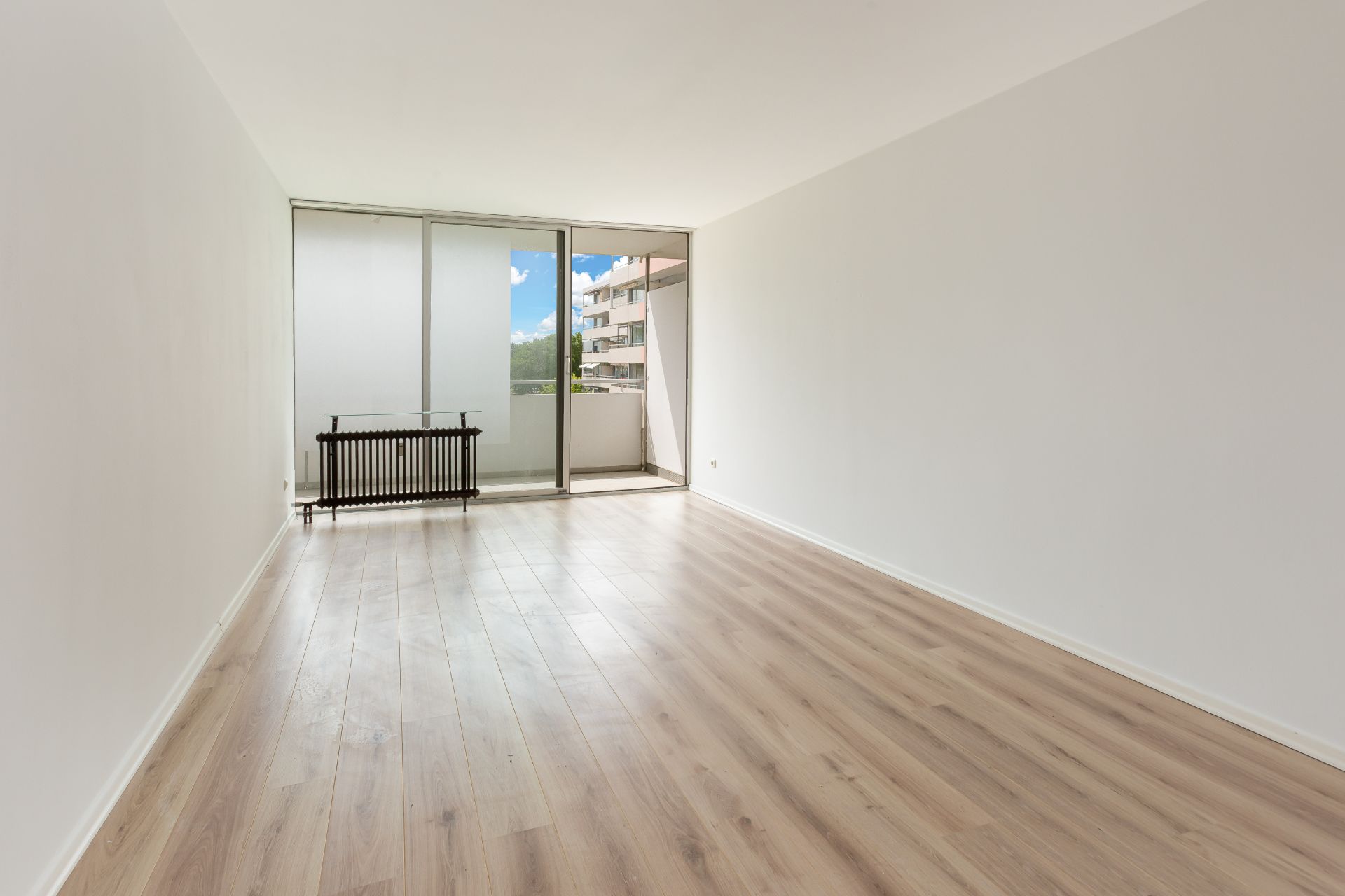 Leeres Wohnzimmer mit Holzboden und großer Fensterfront zu einem Balkon.