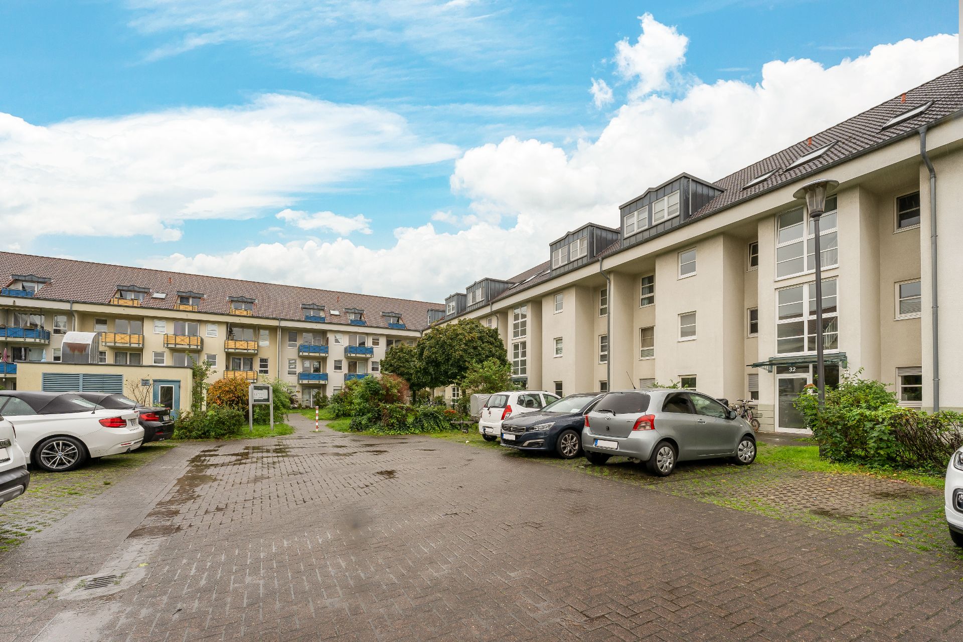 Mehrfamilienhaus mit geparkten Autos auf einem gepflasterten Hof und grünem Bewuchs.