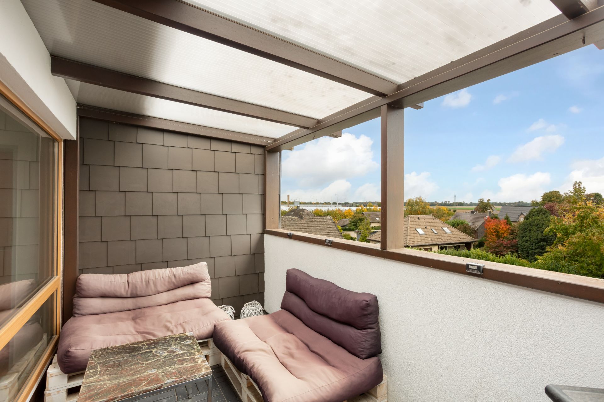 Überdachter Balkon mit zwei gepolsterten Sitzbänken und kleinem Tisch, Blick ins Grüne.