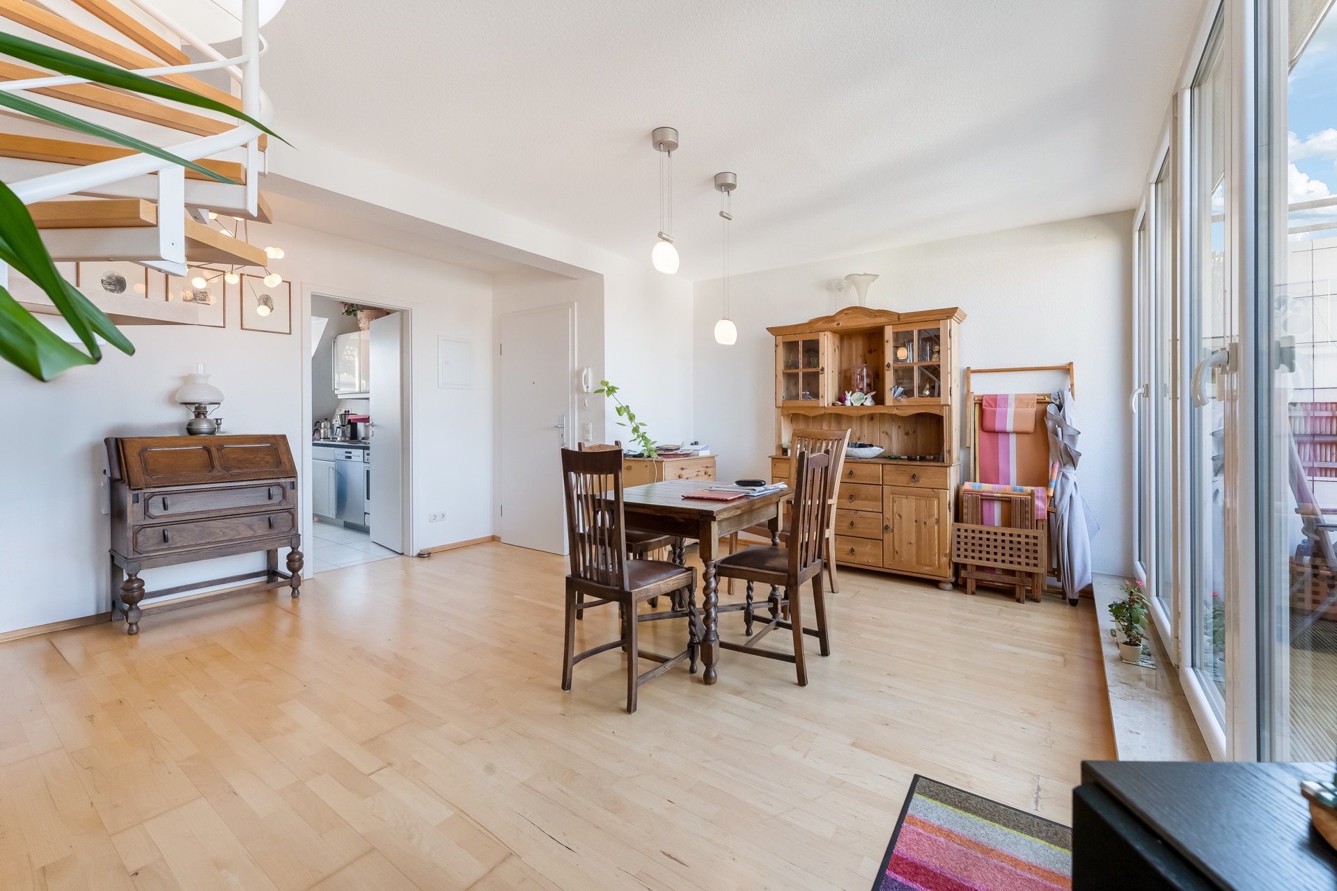Helles Wohnzimmer mit Holzmöbeln, Esstisch, Vitrine, Fensterfront und offener Treppe.