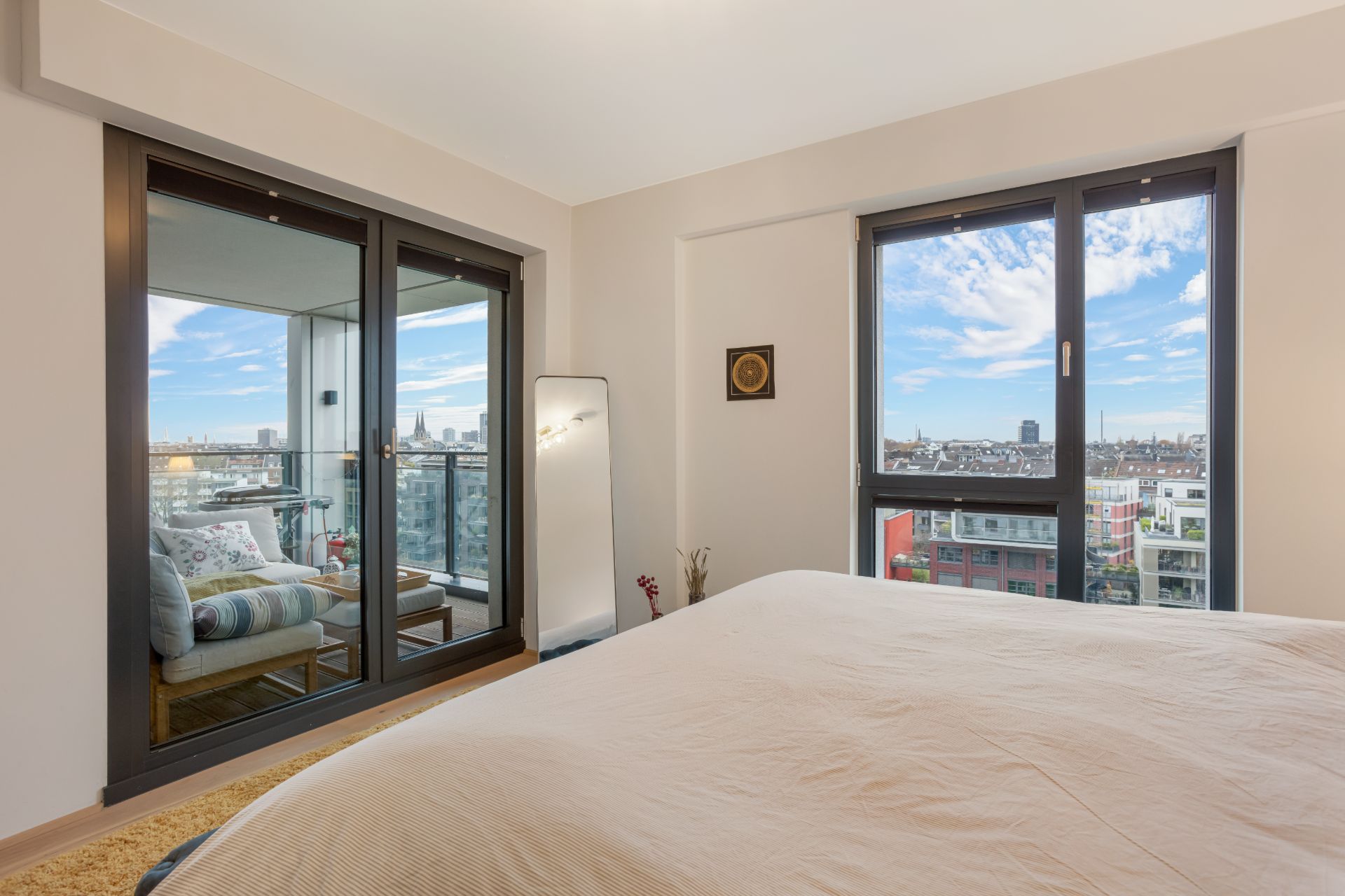 Schlafzimmer mit großem Fenster und Zugang zu einem Balkon mit Sitzgelegenheit, Blick auf die Stadt.