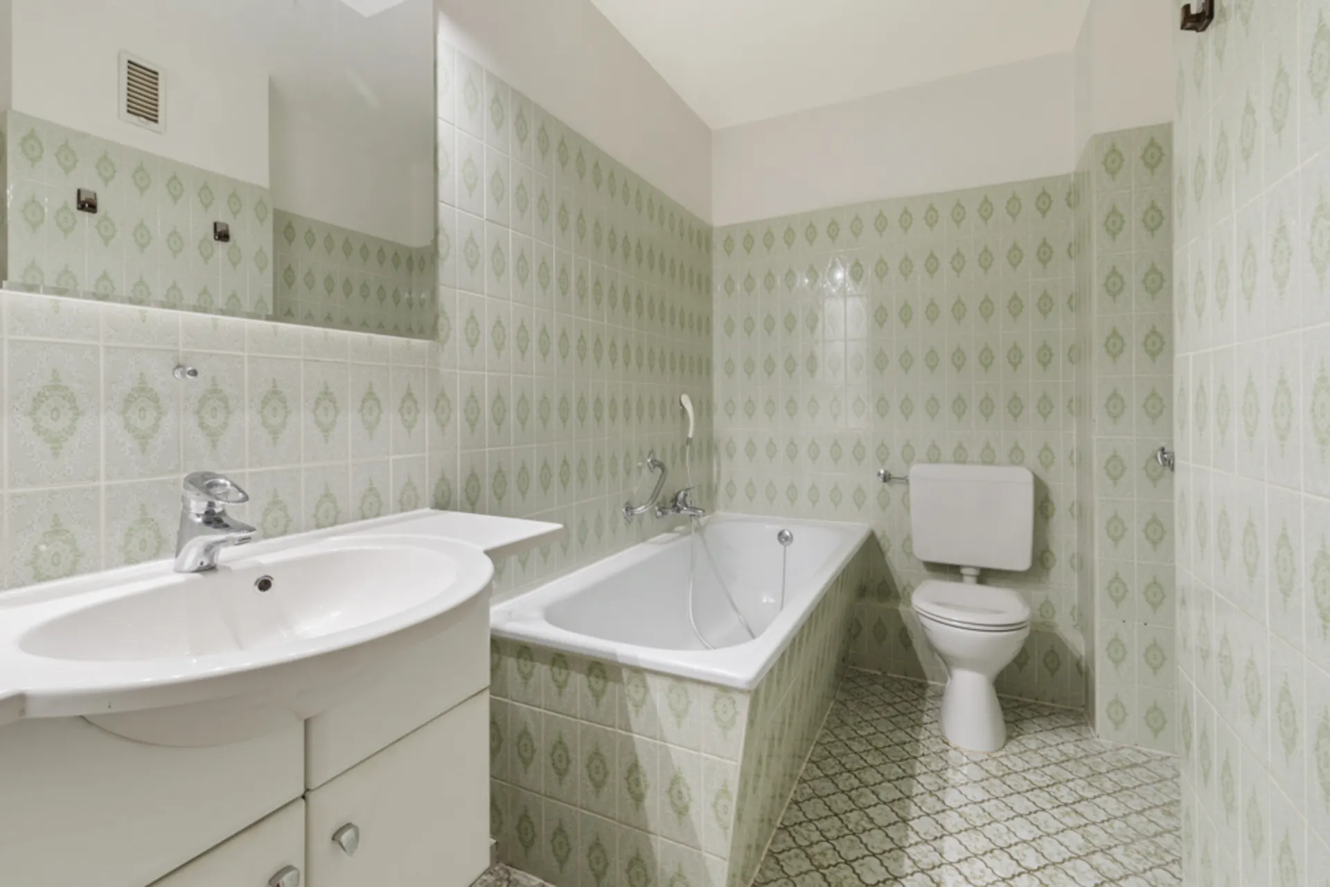 Badezimmer mit Badewanne, Waschbecken und Toilette, grüne gemusterte Fliesen an Wand und Boden.
