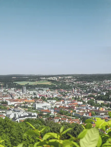 Stadtpanorama mit vielen Gebäuden, Bäumen und grünem Vordergrund bei klarem Himmel.