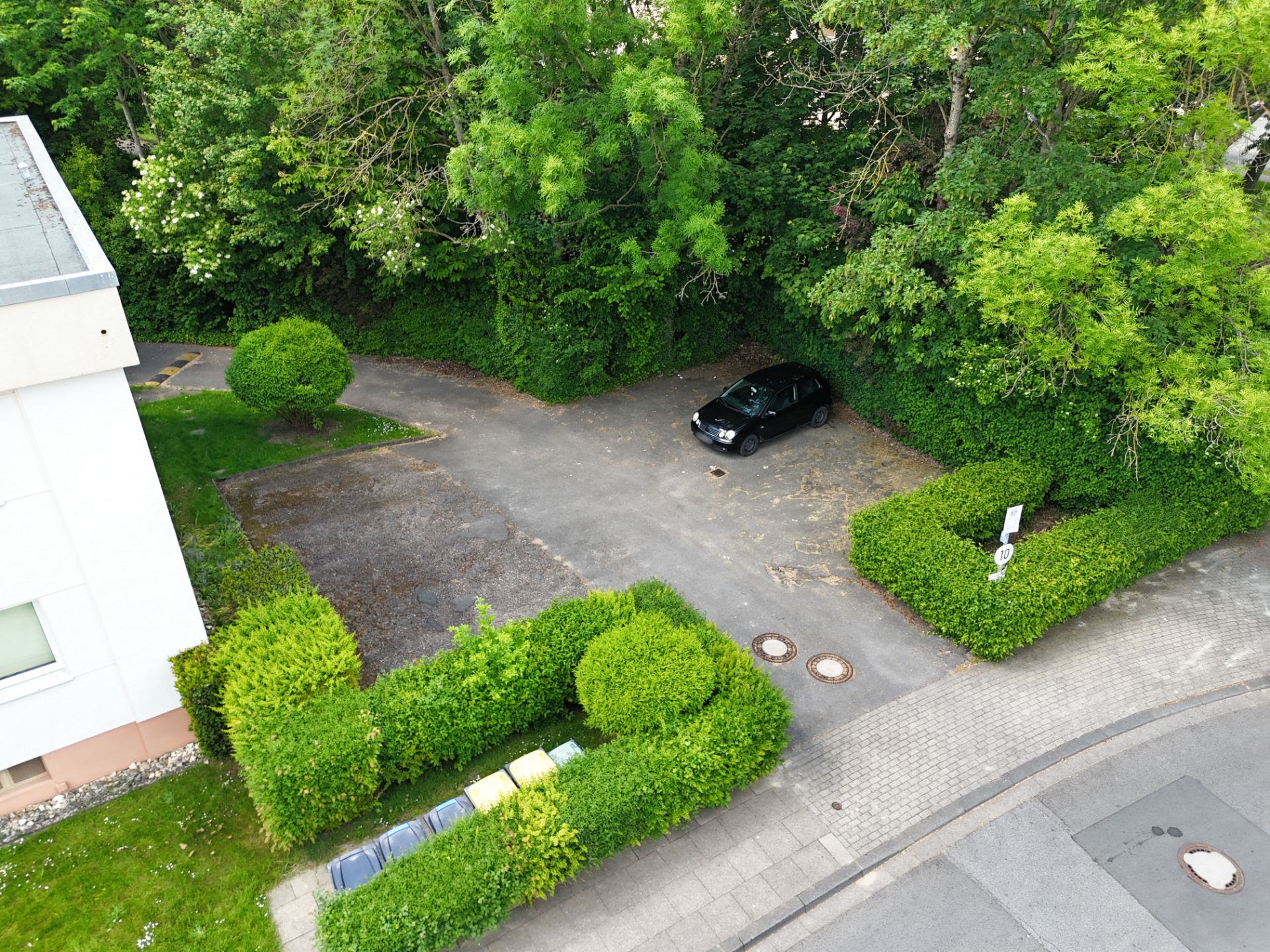 Parkplatz mit einem schwarzen Auto, viel Grün und Mülltonnen neben einem Wohngebäude.