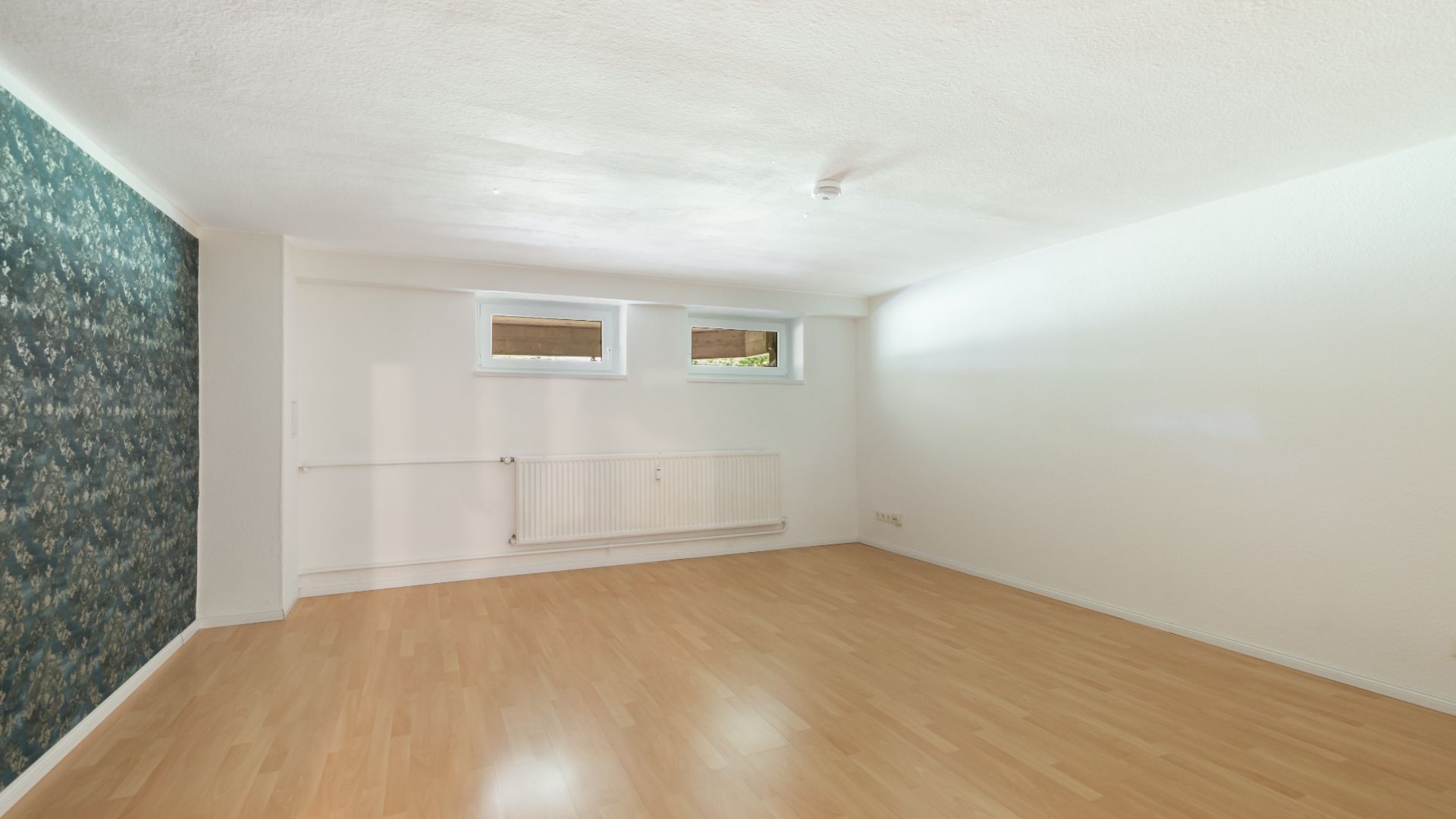 Leerer Raum mit hellem Laminatboden, zwei kleinen Fenstern und einer Wand mit Blumenmuster.