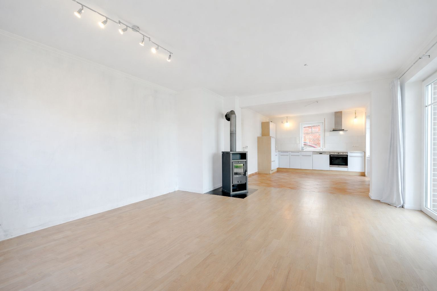 Helles Wohnzimmer mit Holzofen und offener Küche, große Fenster und Parkettboden.