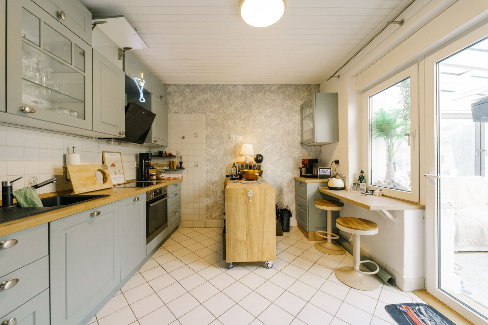 Helle Küche mit grauen Schränken, Küchengeräten, Tresen und zwei Hockern am Fenster.