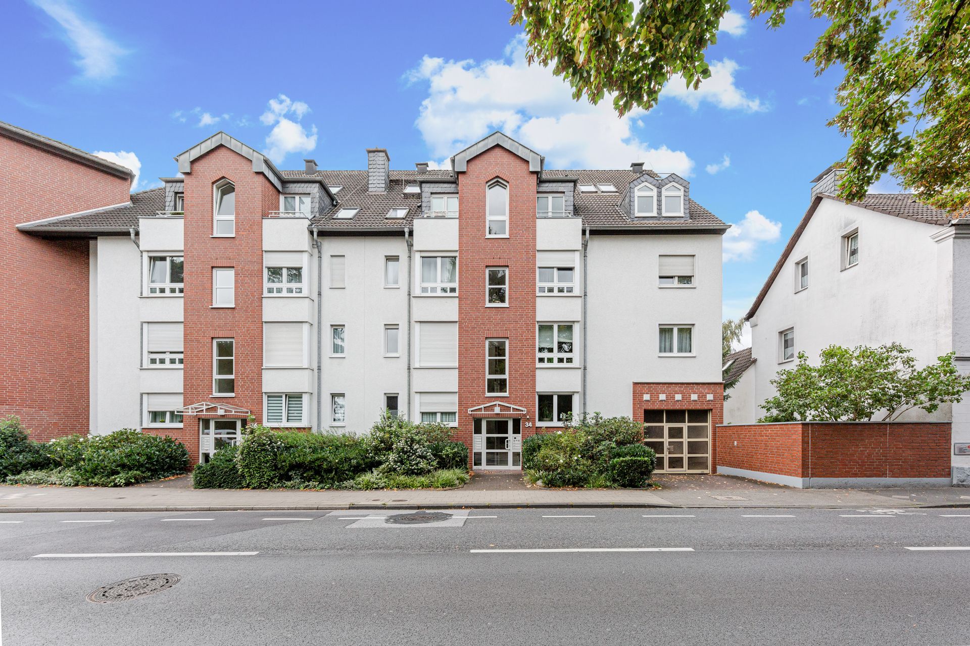 Mehrstöckiges Wohnhaus mit roter und weißer Fassade, Garagentor und begrünter Vorgarten an der Straße.