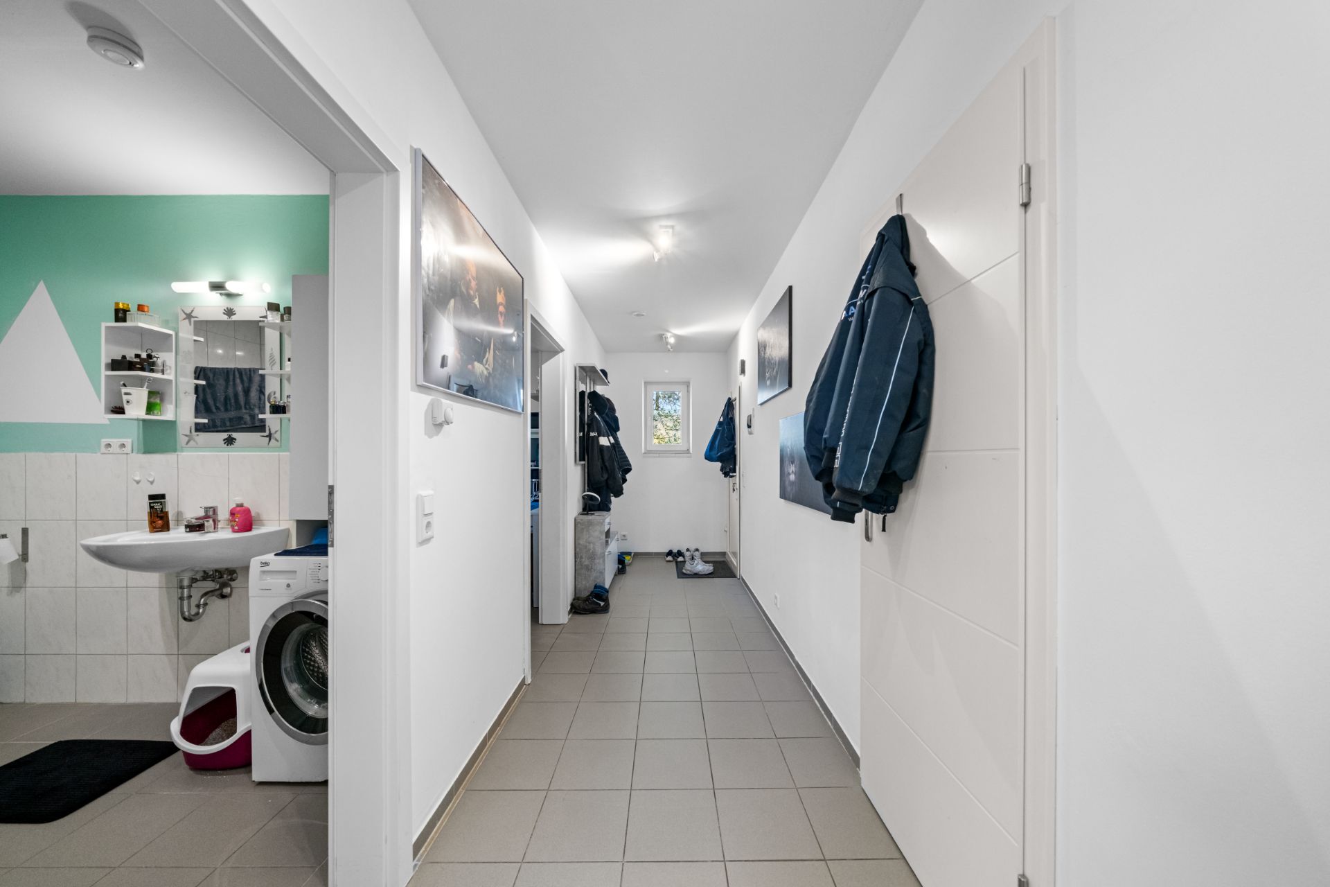 Heller Flur mit gefliestem Boden, Jacken an Haken und Blick ins angrenzende Badezimmer mit Waschmaschine.