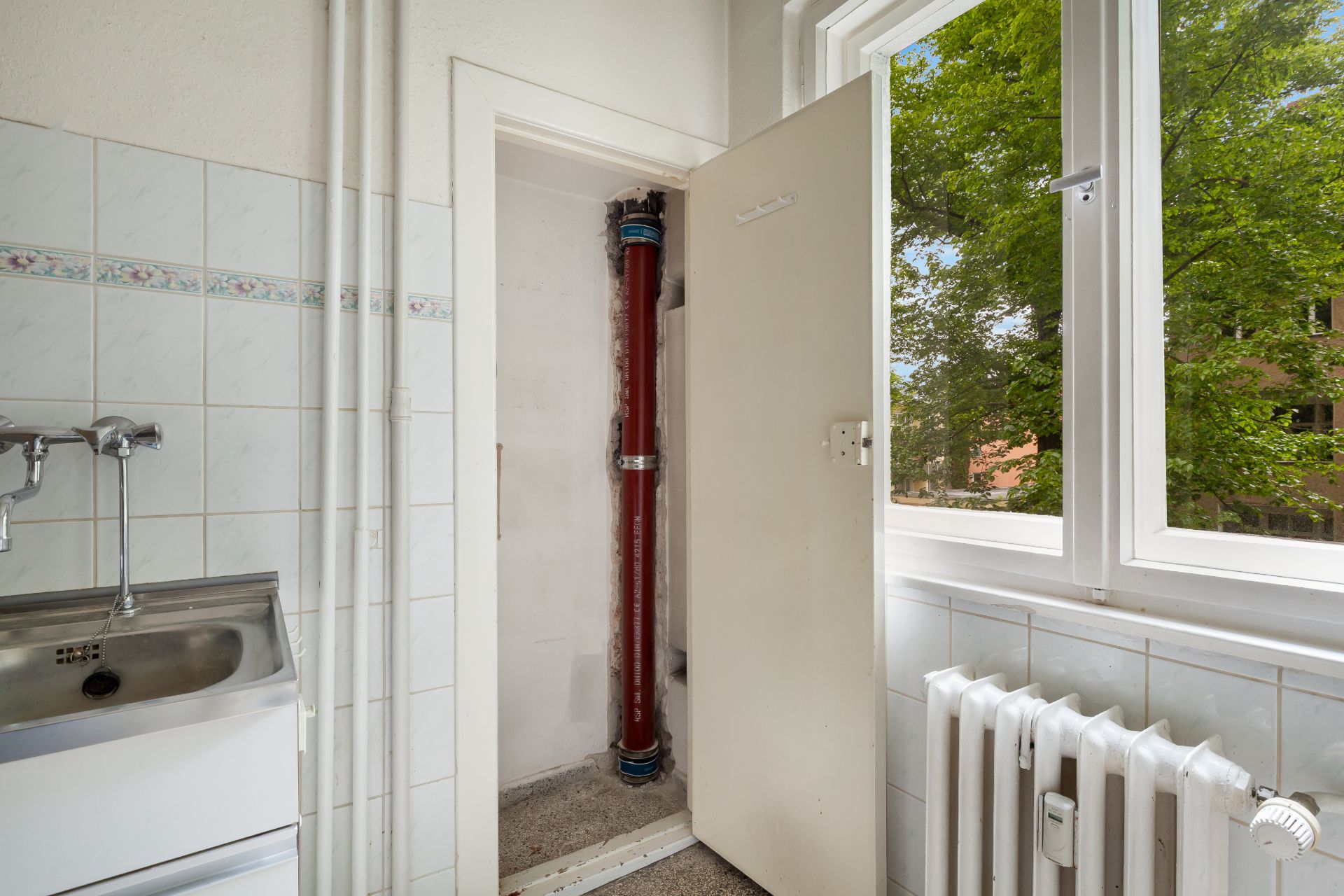 Offener Schrank mit rotem Rohr in einer hellen Küche mit Fenster und Spüle.