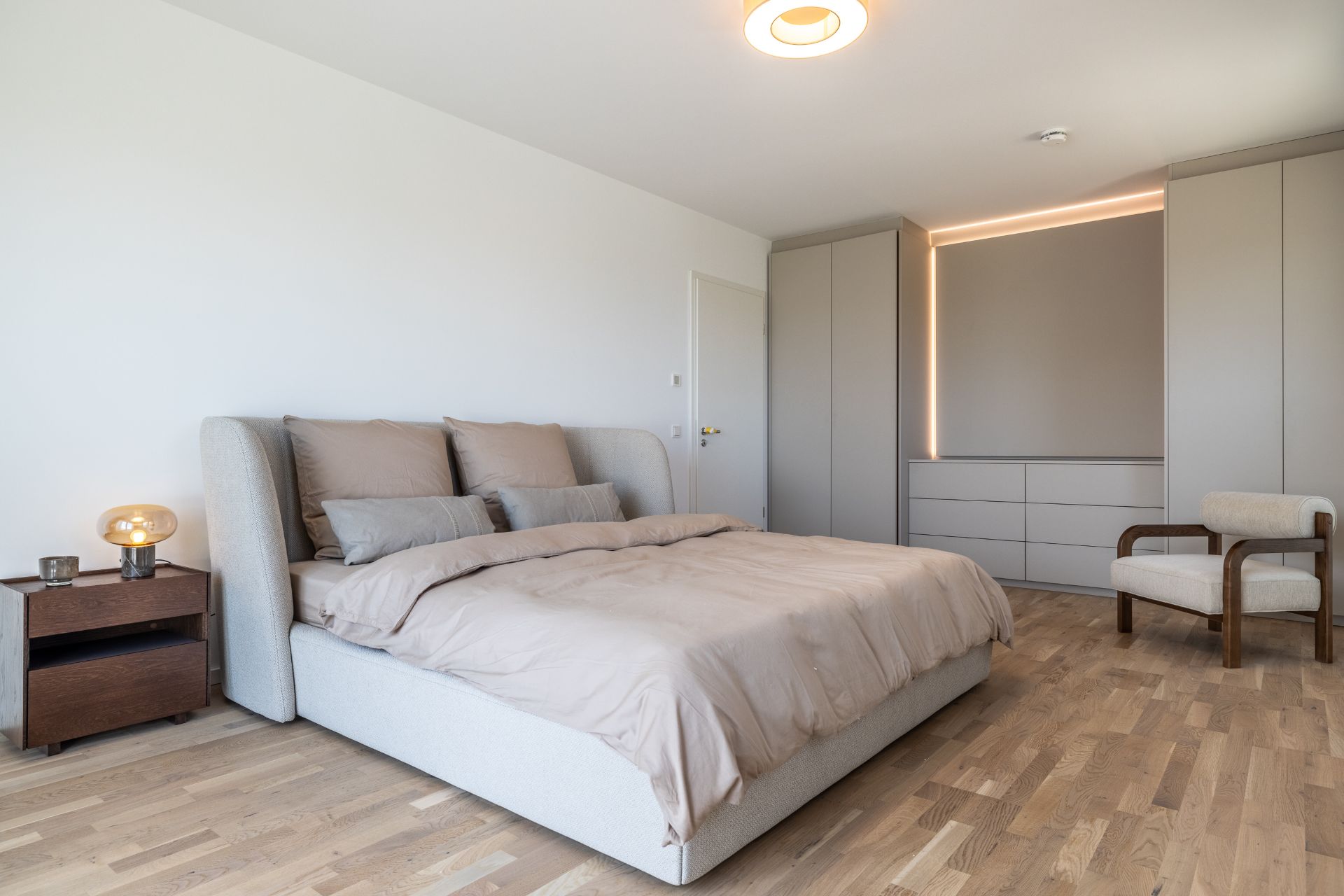 Modernes Schlafzimmer mit großem Bett, Nachttisch, Sessel und Einbauschränken auf Holzfußboden.