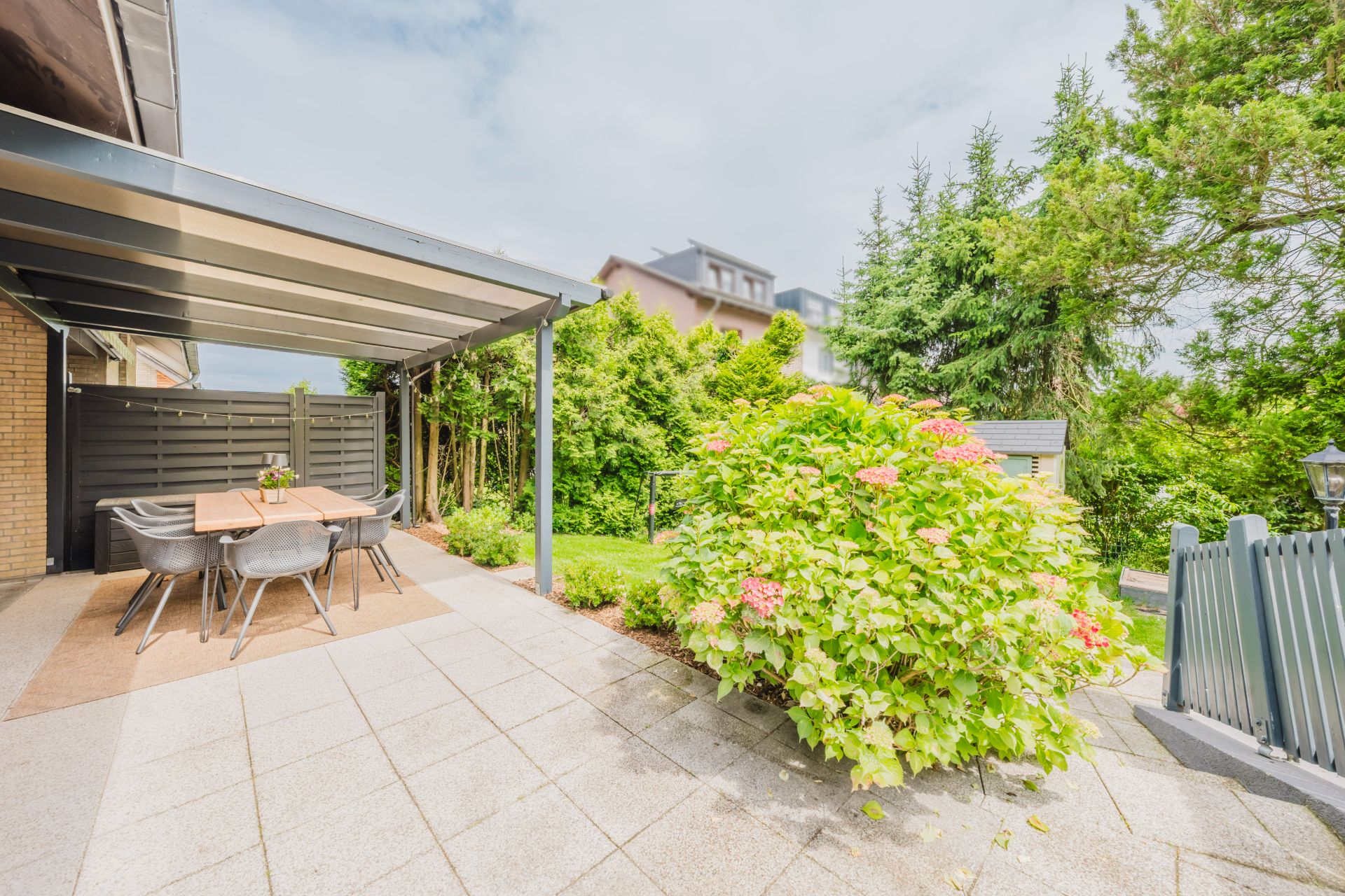 Überdachte Terrasse mit Esstisch und Stühlen, daneben blühender Strauch und Garten.